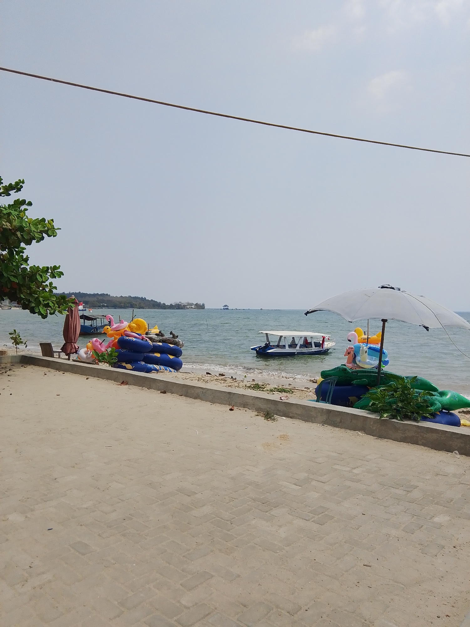 The image depicts a beach scene in Pejagan, Indonesia, under a bright, hazy sky. In the foreground, a paved walkway made of interlocking bricks leads towards the beach. Along the edge of the walkway, a concrete border separates it from the sandy beach.
On the beach, various inflatable pool toys are stacked. These include blue inner tubes, a pink flamingo float, a yellow ring float, and several green crocodile-shaped floats. A large white umbrella is open, providing shade over some of the inflatable toys. A few small green plants are growing along the concrete border.
In the water, a white boat with a covered seating area is visible, with several passengers inside. Another, larger boat, partially obscured, is moored further offshore. The sea is calm and appears a light blue-green. In the distance, a tree-lined coastline with some buildings can be seen. A thin wire runs horizontally across the upper portion of the image. The overall impression is a quiet, sunny day at a tropical beach, likely prepared for recreational activities.