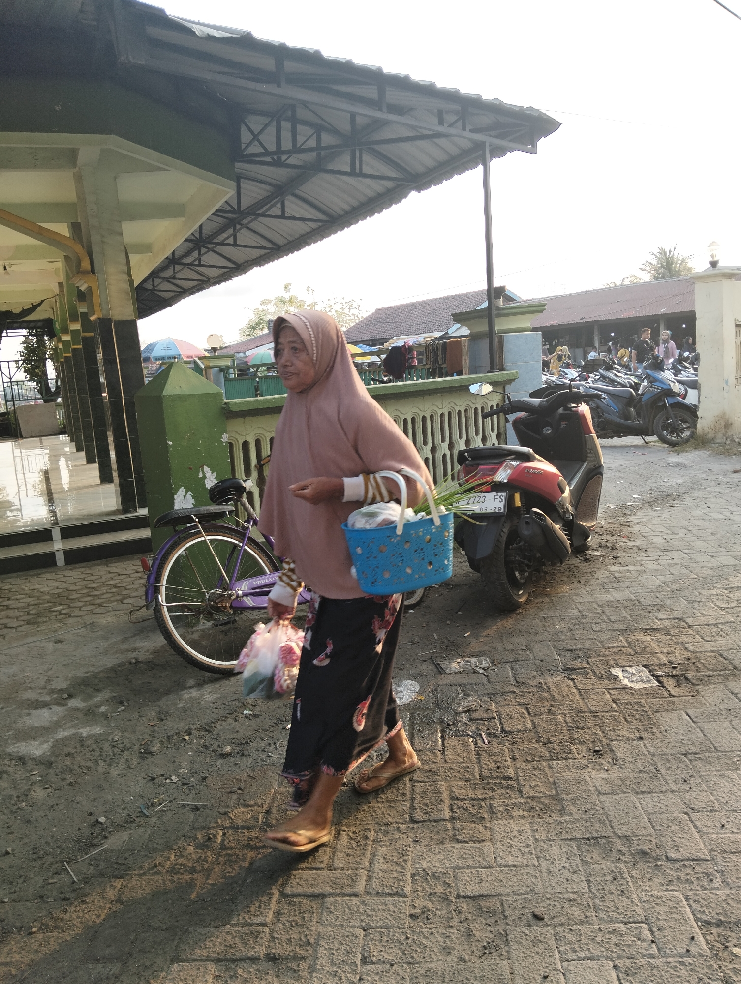In the city of Banjarwati, Indonesia, an elderly woman wearing a pink hijab and a patterned black skirt walks on a paved surface. She is carrying a blue plastic basket filled with greenery and a plastic bag of produce. To her left, a purple bicycle is parked. In the background, several motorcycles are parked, and a building with a corrugated metal roof is visible. The scene appears to be a public area, possibly a market or a street. The lighting suggests it is late afternoon or early evening, with long shadows cast across the ground.