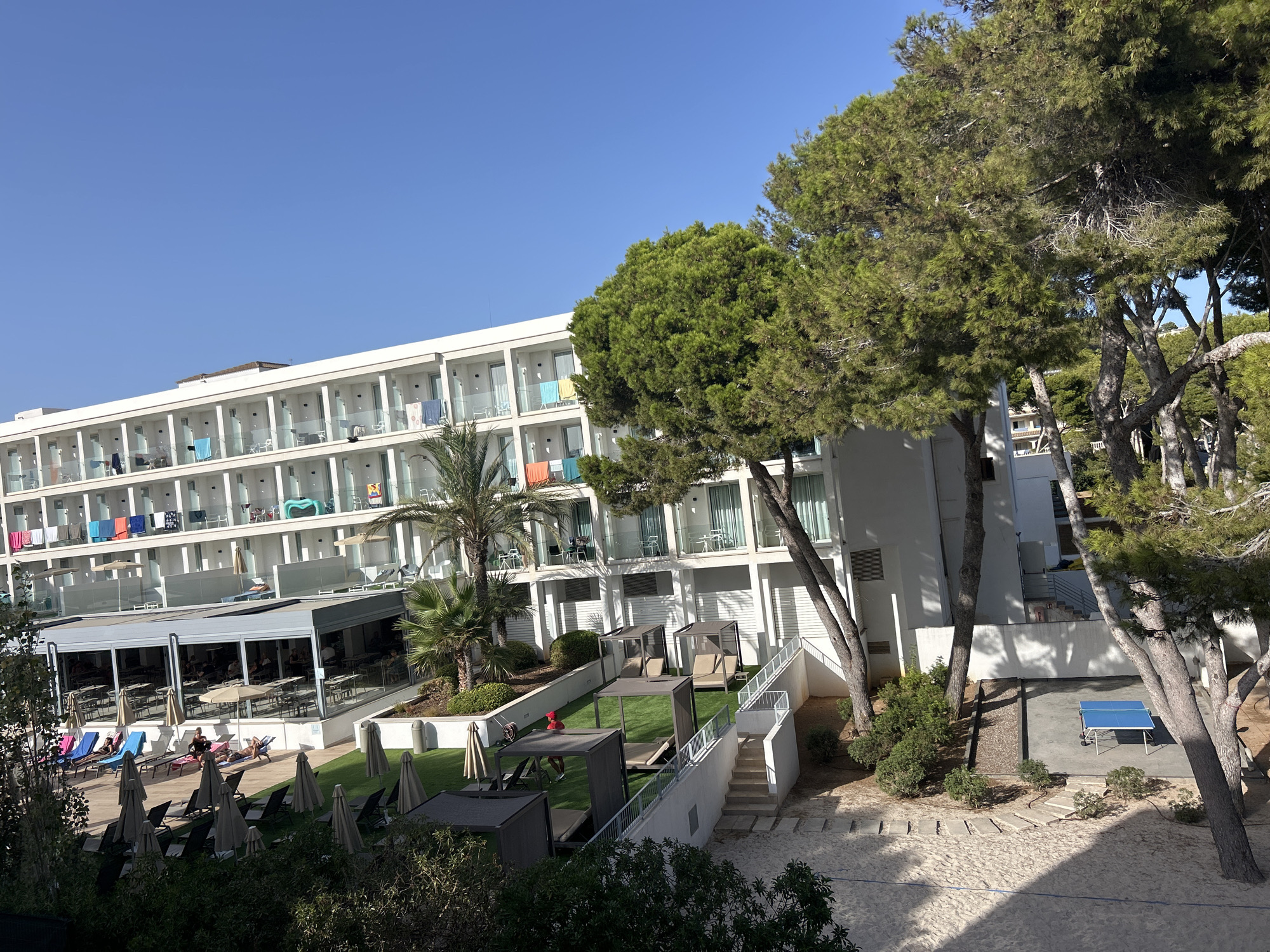 The image shows a multi-story white hotel building with numerous balconies, some of which have towels hanging from them. In front of the hotel, there is a swimming pool area with lounge chairs and umbrellas, as well as a covered dining area with tables and chairs. Several palm trees and large pine trees are visible, some casting shadows on the sand and pavement. There is also a ping pong table in the lower right portion of the image. The scene is outdoors on a sunny day with a clear blue sky. The location is Capdepera, Spain.