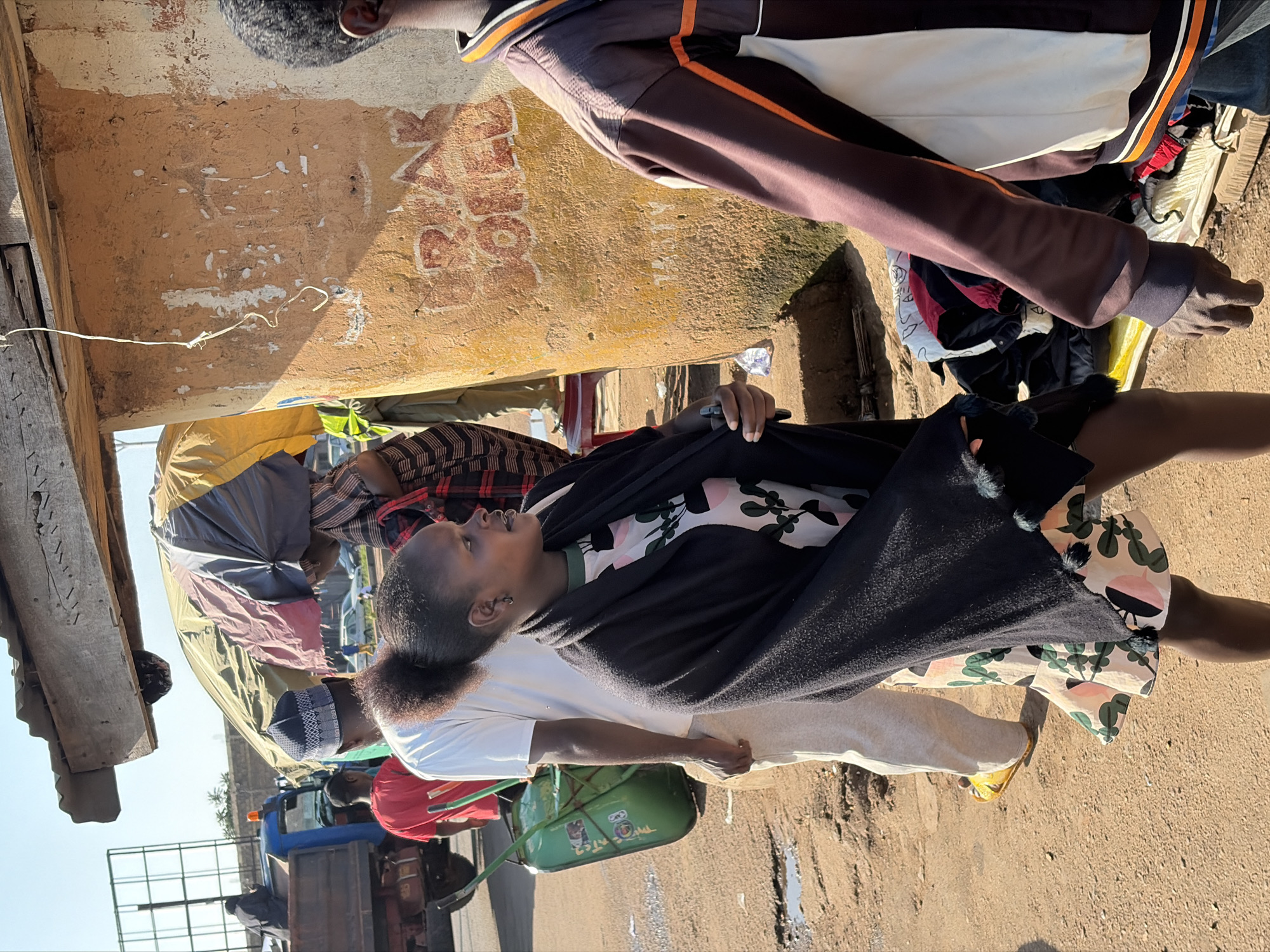 The image depicts an outdoor scene in Gura Top, Nigeria. In the foreground, a young woman with dark hair pulled back in a ponytail is walking. She is wearing a dark shawl over a dress with a floral pattern. To her left, a person in a white t-shirt and light-colored pants is pushing a green wheelbarrow. Behind them, a man in a red t-shirt is also present. In the background, a blue truck is parked. The environment is dusty and appears to be a market or roadside area, with structures made of wood and mud. There are also some items of clothing and other goods laid out. The sun is shining, casting shadows.