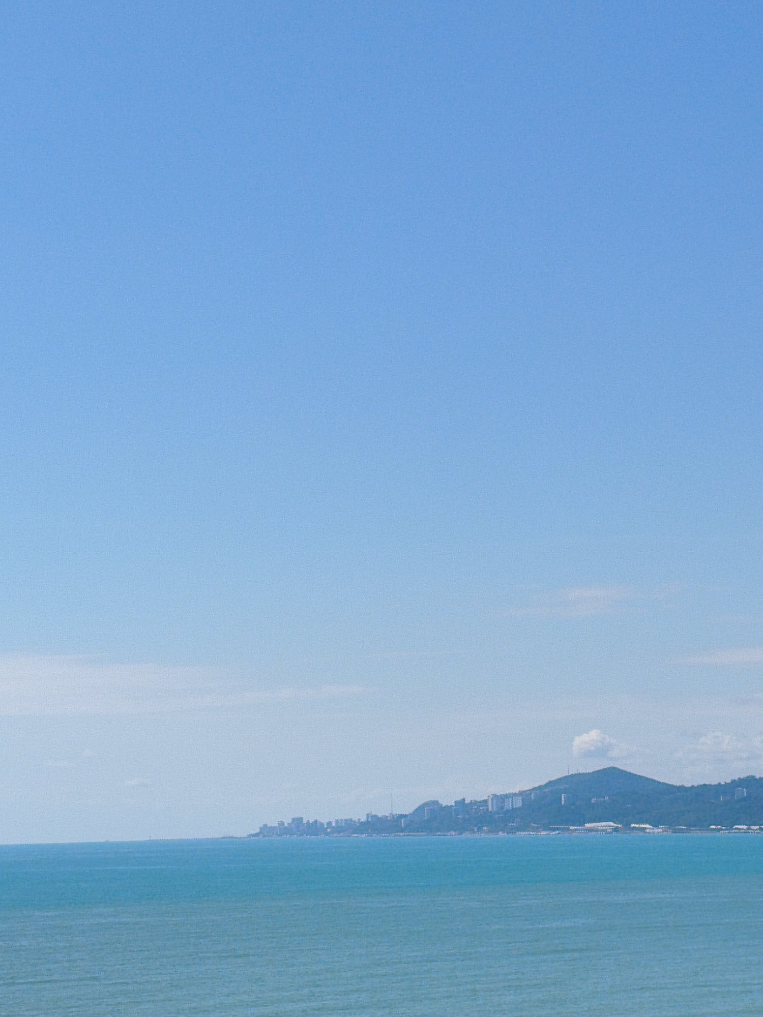 The image captures a panoramic view of the Black Sea coast in Sochi, Russia, on a clear, sunny day. The expansive, bright blue sky dominates the upper portion of the frame, with a faint, wispy cloud formation visible on the horizon. Below the sky, the turquoise waters of the Black Sea stretch across the foreground, exhibiting a slight ripple texture. Along the distant coastline, a chain of buildings, indicative of a city, is nestled against the backdrop of a lush, green, mountainous landscape. The buildings appear to be a mix of modern architecture and older structures, with some taller buildings interspersed among lower-rise developments. The natural topography of the mountains is pronounced, with slopes covered in dense vegetation. A small, puffy white cloud hovers over the peak of one of the mountains. The scene suggests a typical coastal resort area with a blend of urban development and natural beauty.