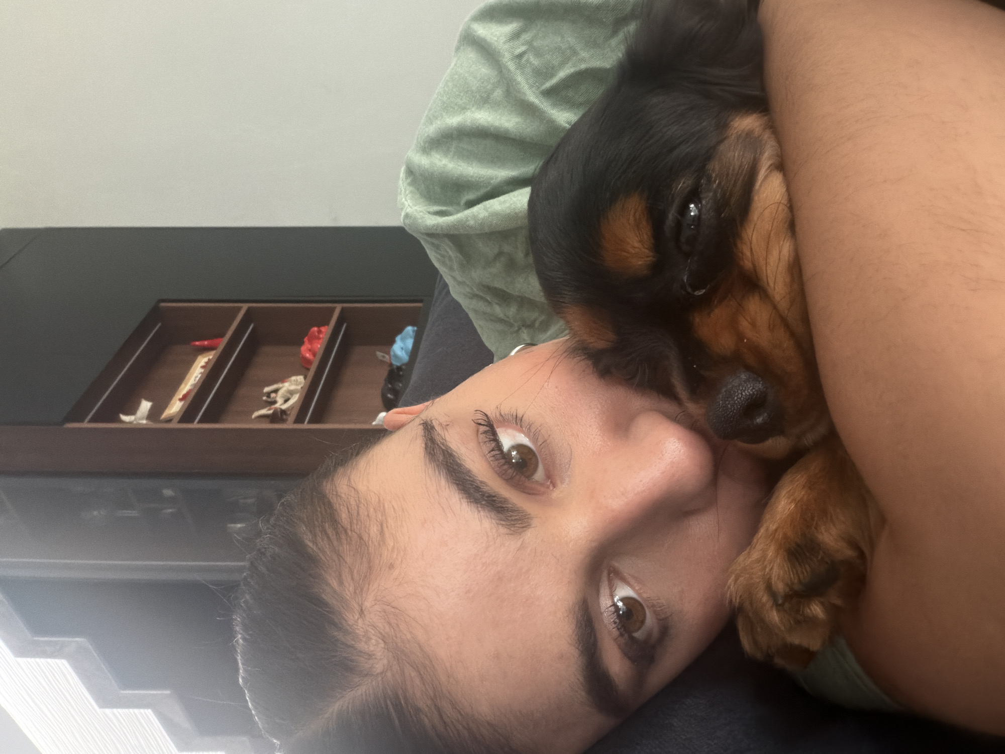 A woman is lying down, holding a small dog in her arms. The dog is mostly black with brown markings on its face and paws. The woman has brown eyes and dark hair. Behind the woman and the dog is a green fabric, possibly a shirt or pillow. In the background, there's a dark brown wooden unit with several compartments containing various small items. The lighting is bright and the image appears to be taken indoors. There is no verifiable evidence to link this picture with Puebla, Mexico.
