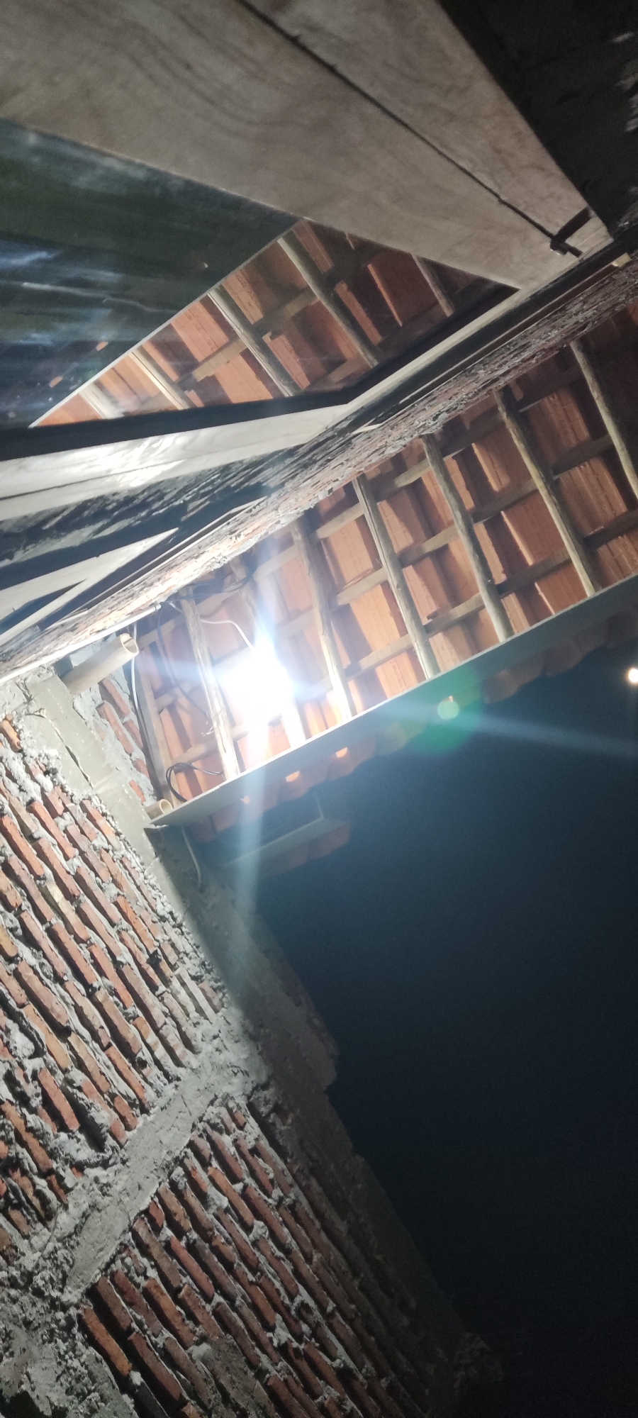 The image is an upward-facing shot of an unfinished structure, likely under construction. The foreground features a brick wall with visible mortar, constructed in a staggered pattern. Above the wall, wooden beams and terracotta roof tiles are visible, forming the roof structure. A bright light bulb illuminates the scene from within the structure, casting a strong glare and lens flare. The background is dark, indicating nighttime. The scene is consistent with construction activity in a residential area, and the presence of unfinished brickwork and roof trusses suggests an ongoing building project. This view is characteristic of the many developing residential areas found in Ciledug, Indonesia, where such construction is a common sight.