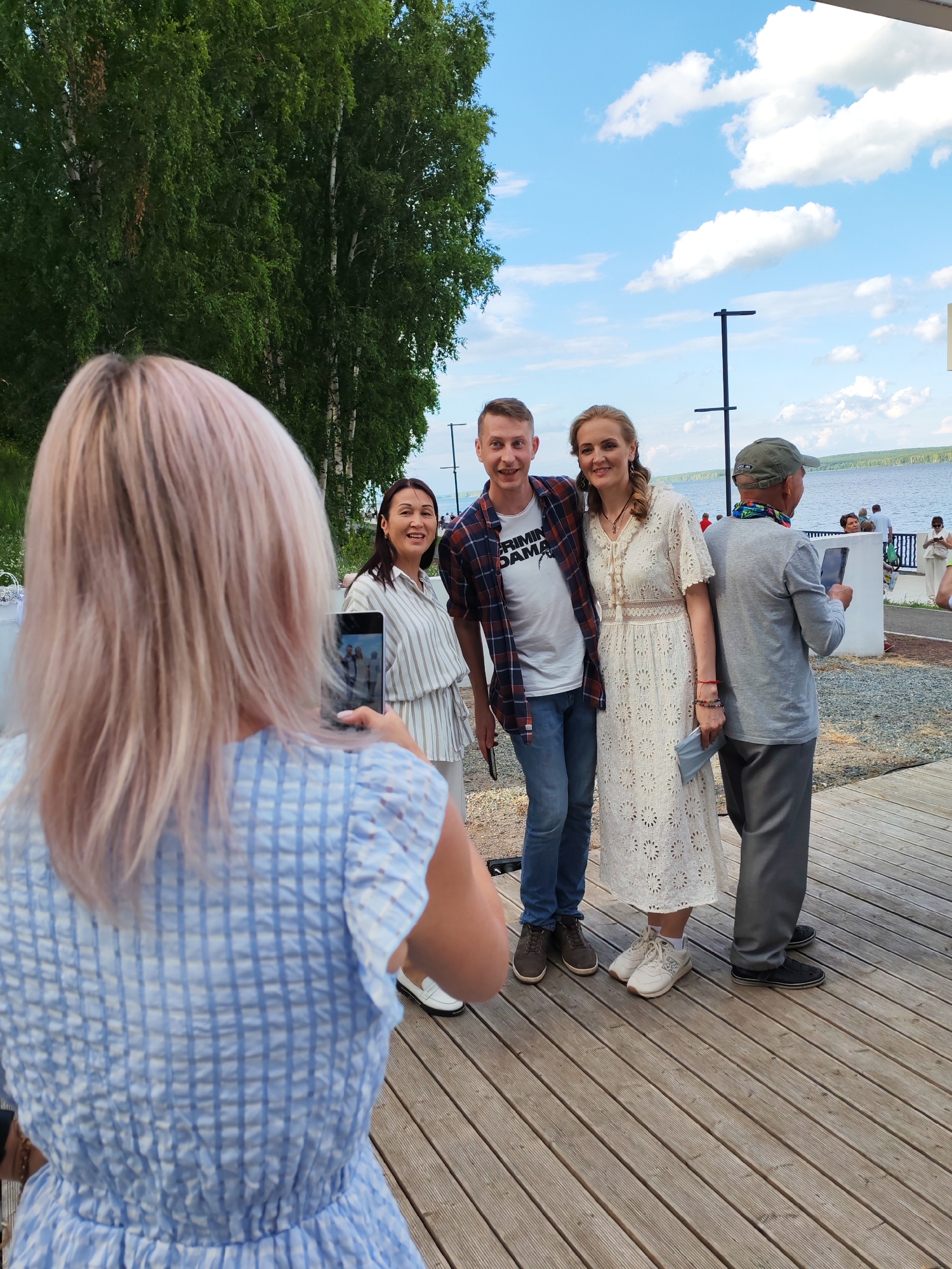 The image shows a group of people posing for a photo on a wooden boardwalk by a body of water, possibly the Kama River, in Chaikovsky, Russia. In the foreground, a woman with blonde hair, wearing a blue and white checkered dress, is taking a photo with her phone of three individuals standing in the middle ground. These individuals include a man in a plaid shirt and jeans, a woman in a white lace dress, and an older man wearing a grey shirt, grey pants, and a green cap. The background features trees, a cloudy sky, and other people in the distance. The overall scene appears to be a casual outdoor gathering.