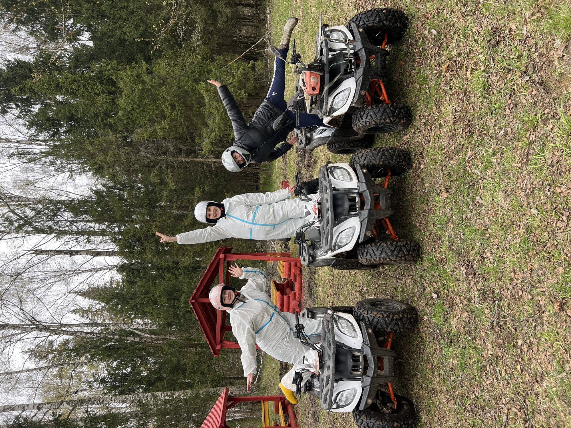 Three people are on ATVs in a grassy area near a forest. The person on the top ATV is wearing a black jacket and pants and a white helmet. The two people on the bottom ATVs are wearing white protective suits and helmets. The ATVs are white and gray with orange accents. A red and yellow structure is visible in the background. The location may be near the city of Ширяево, Russia, based on the geographical location of this picture, which might be a suburb or rural area.