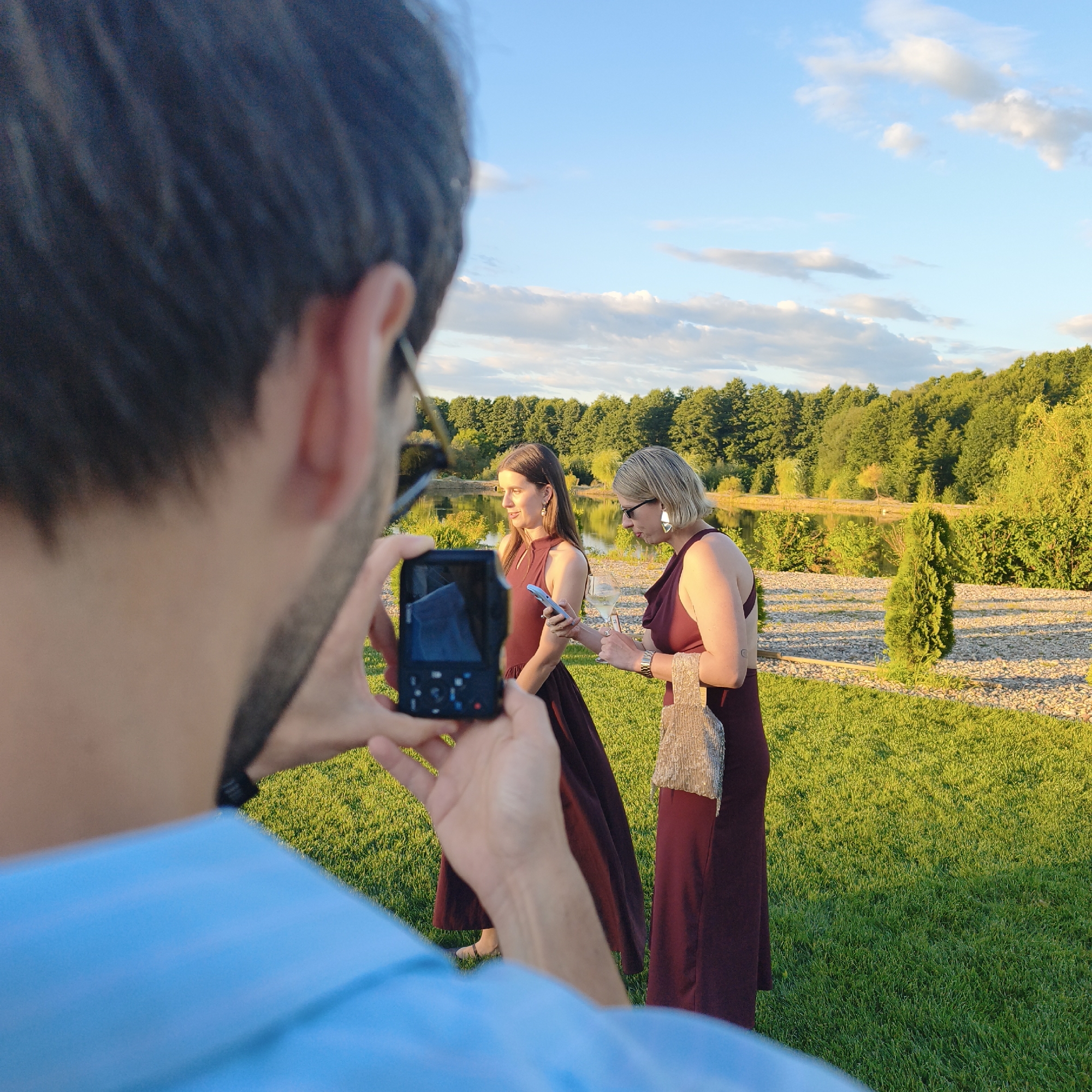 In the foreground, a person's back and shoulder, clad in a light blue shirt, are visible as they hold a compact digital camera to take a photograph. The camera is pointed towards two women standing in a grassy outdoor area. The woman on the left, with long brown hair, is wearing a dark red sleeveless dress and is looking to her left. She is holding a smartphone in her right hand. The woman on the right, with short blonde hair, is wearing a similar dark red sleeveless dress and large silver earrings. She is wearing glasses and is looking down at something in her hands, possibly a wine glass. She is also holding a small, sparkly clutch bag.
The background features a body of water, likely a lake, with a treeline of lush green trees bordering it. The sky is a clear blue with scattered white clouds, suggesting it is late afternoon or early evening. The ground is covered in bright green grass, with patches of gravel and some small evergreen trees. The location appears to be a park or a scenic outdoor venue. The overall atmosphere is one of a social gathering or event taking place in a natural setting.