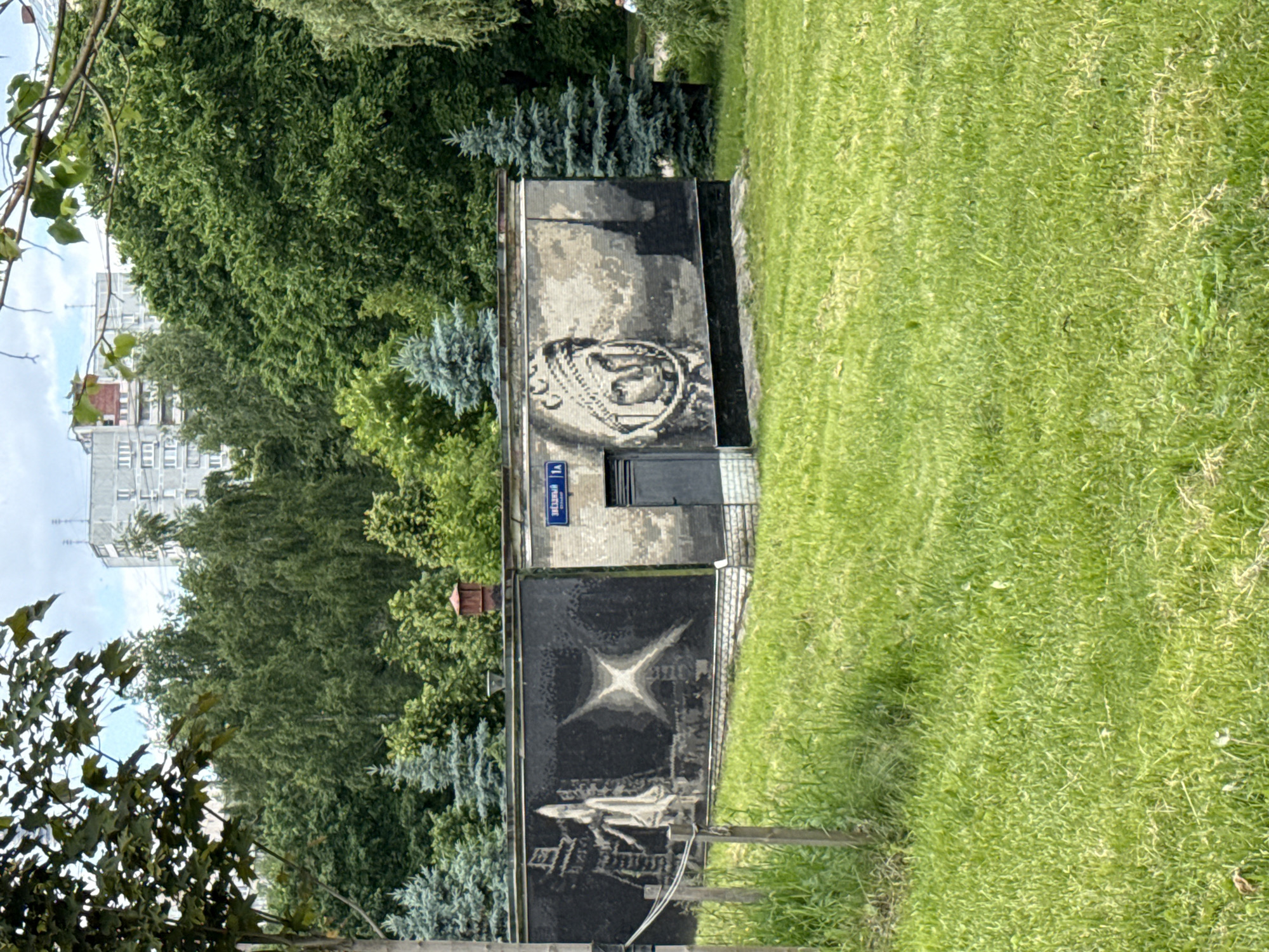This outdoor photograph captures a small, rectangular concrete building adorned with two distinct murals, set within a verdant urban park or green space. The image appears to be taken during daylight hours under a partly cloudy sky.
On the right side of the building, a large circular medallion-style mural depicts the portrait of a person, possibly a cosmonaut, wearing a distinctive helmet or headgear. Below and to the left of this mural, a small blue street sign is affixed to the building, displaying white Cyrillic text that reads "улица" (ulitsa, meaning street) and likely "1А" or "10". A dark-colored door is also visible on this side of the structure.
The left side of the building features another mural with a space theme. At the top, a prominent stylized star or celestial body with radiating points is depicted. Below it, a detailed representation of a space shuttle, possibly the Buran, is shown on a launchpad.
The building is surrounded by neatly kept green grass. In the background, dense foliage of various trees and bushes, including some evergreen conifers, creates a natural backdrop. A multi-story residential building with numerous windows can be seen in the far distance, partially obscured by the trees. No people or specific activities are visible in the scene. The murals, especially the space shuttle, suggest a commemoration of space exploration, fitting the location context of Moscow, Russia.