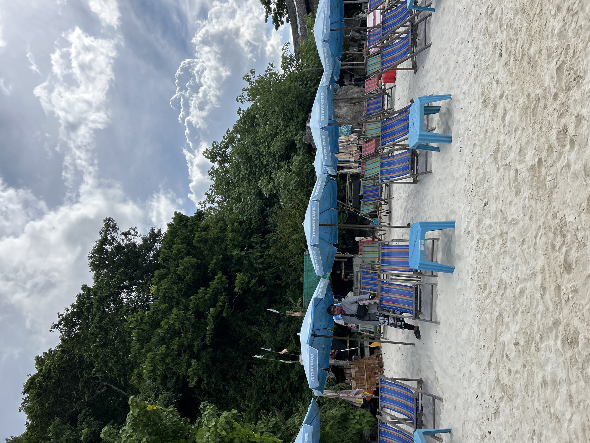 The image depicts a beach scene in Tambon Phru Nai, Thailand. In the foreground, rows of empty, colorful striped lounge chairs and small blue plastic tables are arranged on white sand. Behind the chairs, several light blue umbrellas with the text "JETSKI CLUB" are set up. A person, wearing a grey jacket, patterned pants, a pink scarf, and sunglasses, is standing near one of the umbrellas, holding a bag and an orange paddle. In the background, lush green trees and foliage cover a hill, and a cloudy sky with patches of blue dominates the upper portion of the image. A small structure, possibly a beach shack or bar, is visible behind the umbrellas.