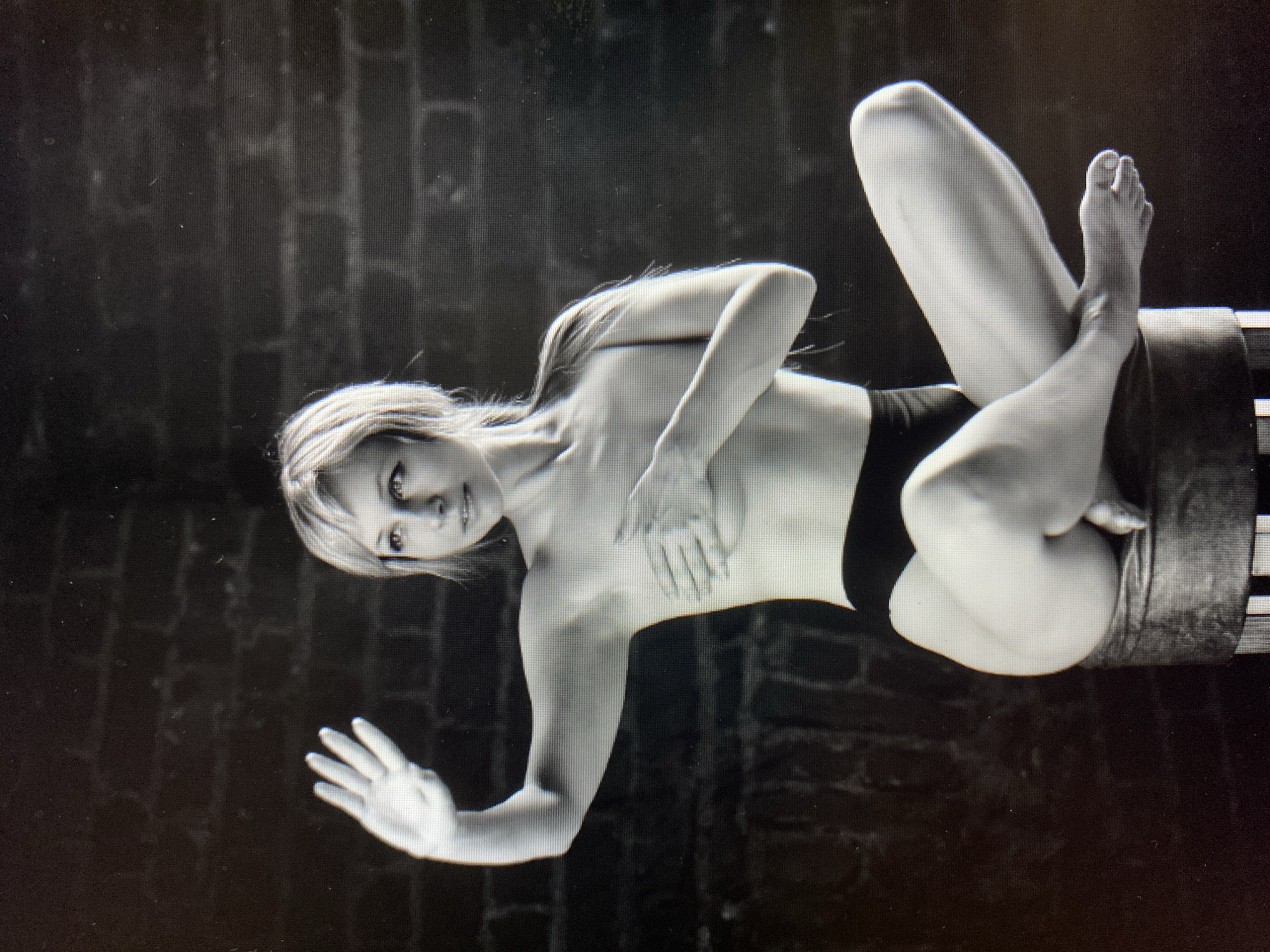 A black and white, full-body studio portrait shows a woman seated cross-legged on a dark, cylindrical stool against a dark, brick-patterned background. The woman has blonde hair and is wearing black underwear. Her left arm is raised with her palm facing forward, and her right hand is placed over her chest. Her legs are crossed, with her right leg over her left. The lighting is dramatic, with strong highlights and shadows.