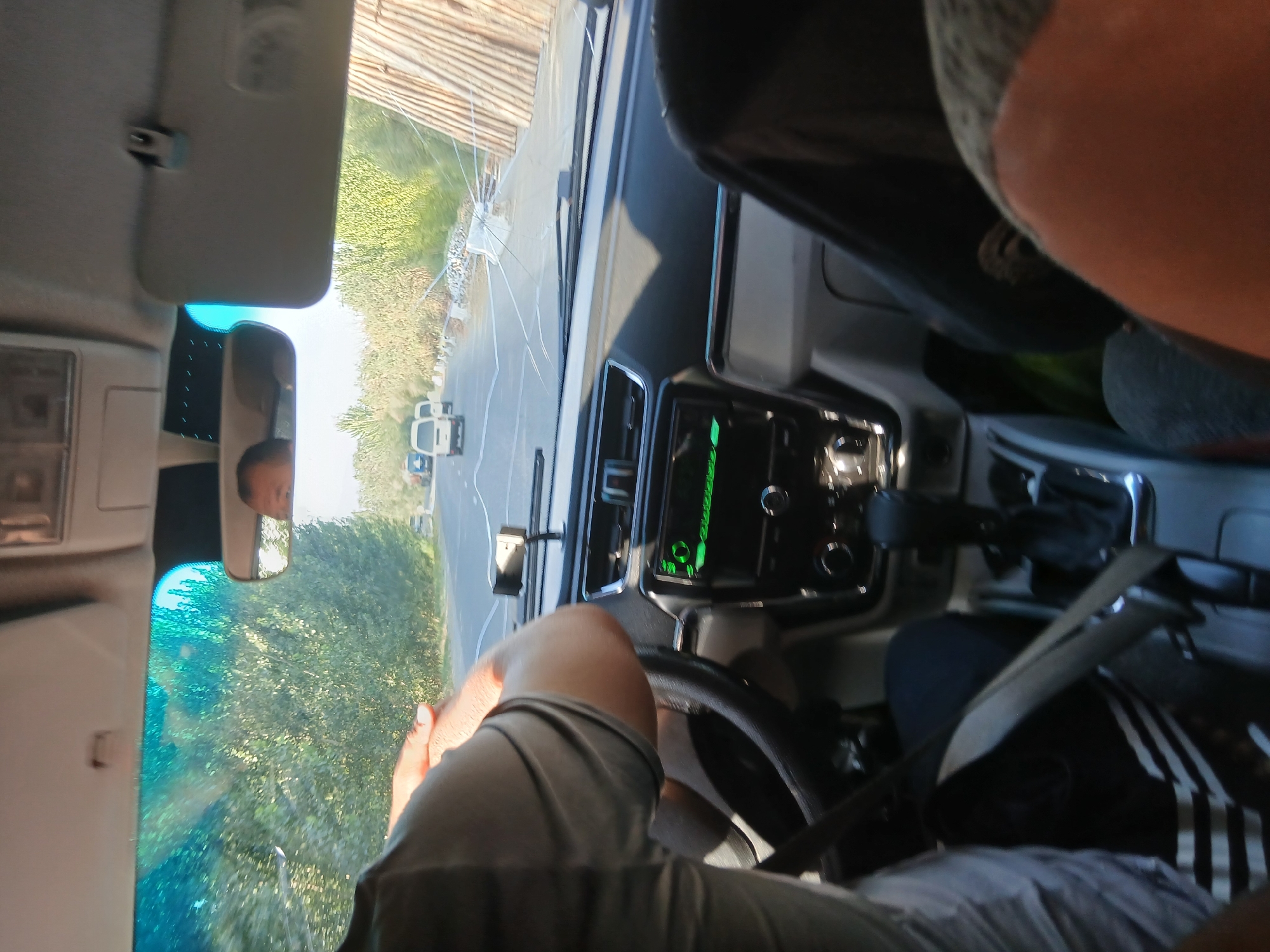 The image depicts the interior of a vehicle, looking forward through the windshield. The driver's left arm and shoulder are visible, resting on the steering wheel, which is partially obscured. The dashboard features a car stereo with a green display, a gear shift, and various controls. Outside the vehicle, a road is visible with a few white vans driving ahead. Trees line the road, and on the right side, there is a pile of wooden poles. The windshield shows a spiderweb crack. The overall scene suggests a journey in progress through a rural or semi-rural area, possibly Khankiz, Uzbekistan, given the context provided. The lighting indicates daytime.