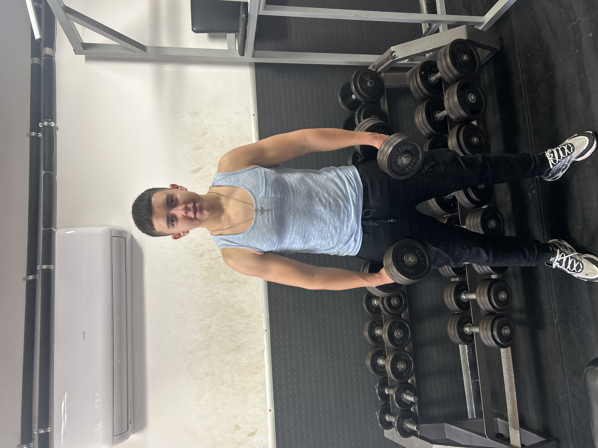 A young man in a light blue tank top and black pants stands in a gym in Zlatograd, Bulgaria, holding dumbbells. He is facing the camera. He has short dark hair. He's wearing white and black sneakers. To his right are racks of dumbbells. A white air conditioning unit is visible on the wall to his left. The floor appears to be black rubber. The image is rotated 90 degrees clockwise.