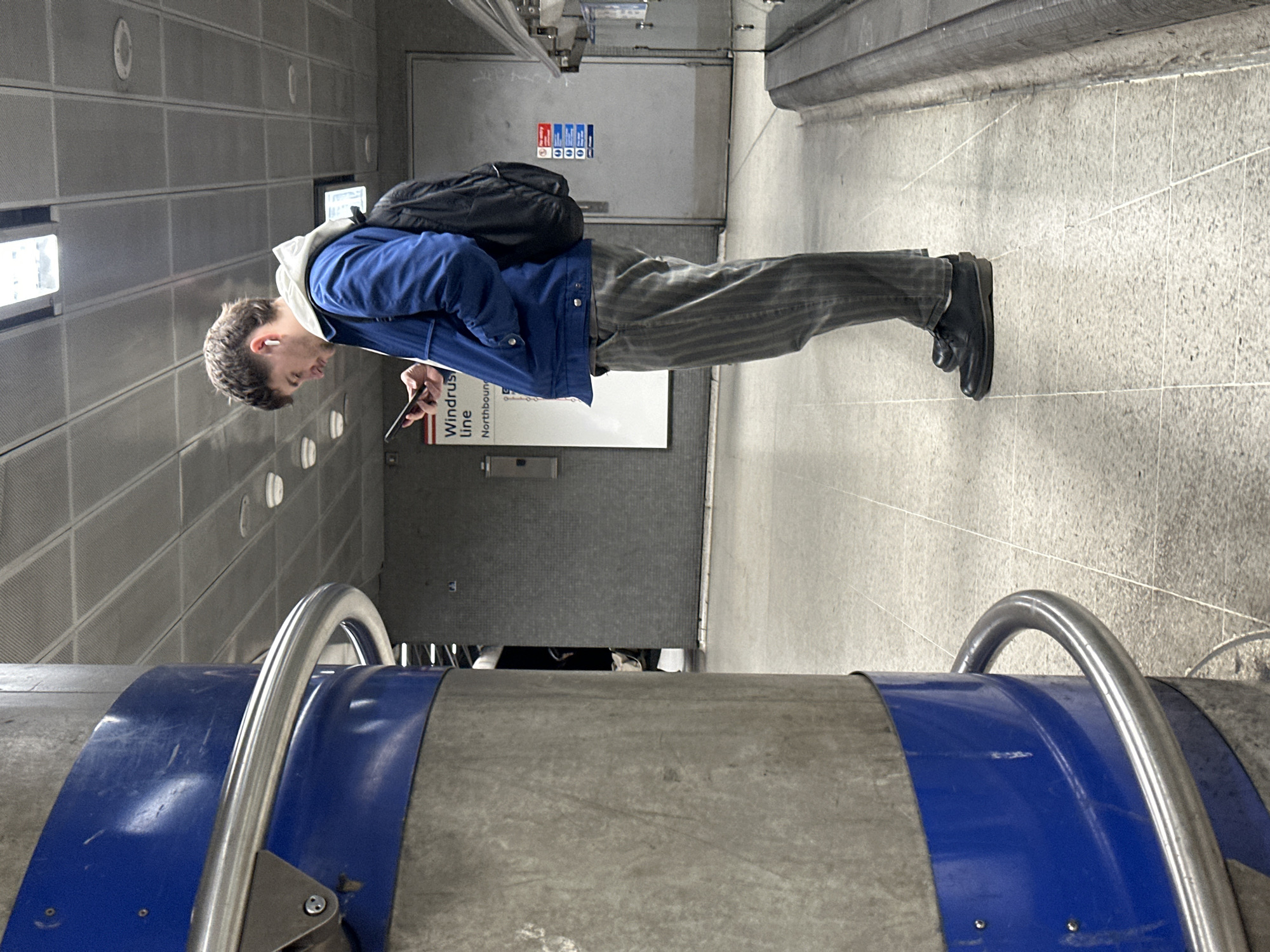 A young adult male stands facing left in an underground station on the Elizabeth Line in London, United Kingdom. He is observed from a slightly low angle, positioned between a concrete pillar wrapped with blue trim and a platform edge. The individual wears a blue jacket over a white hoodie, grey striped trousers, and black shoes. He has a black backpack on his back and white wireless earbuds in both ears. His gaze is directed downwards at a black smartphone he holds in both hands, actively engaging with the device.
To the left, a large concrete pillar with a blue circumferential band and a silver metal handrail occupies the immediate foreground. The station floor is light grey tiled, featuring a grid pattern. In the mid-ground, behind the individual, a white rectangular sign reads "Windrush Line Northbound," indicating the train line and direction of travel. Further right, a grey door with "No entry" and other warning symbols is visible. The background consists of grey tiled walls and an overhead grid ceiling with integrated lighting panels. Artificial light illuminates the scene evenly.