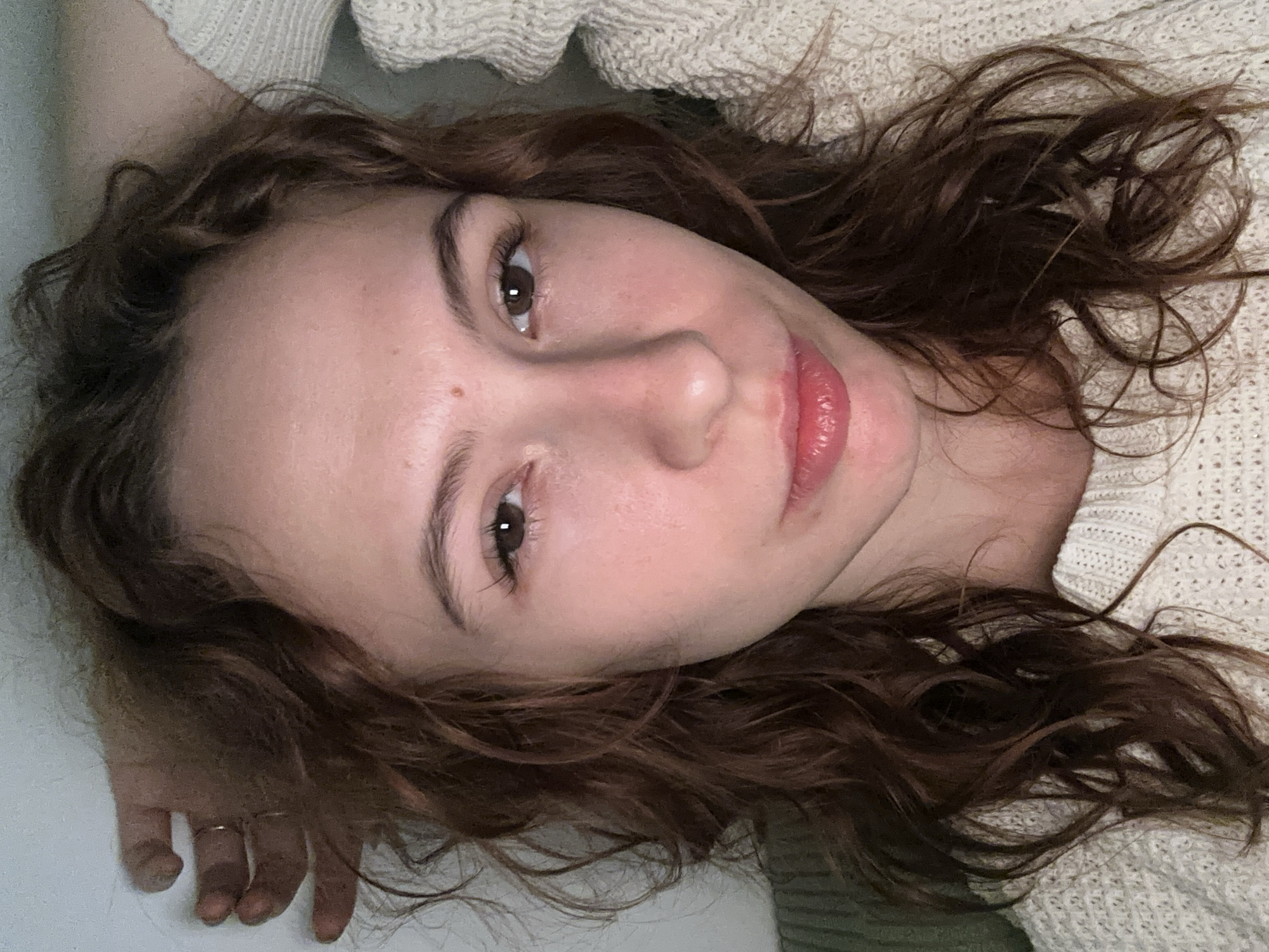 This close-up portrait features a young woman lying down, looking directly towards the camera with a soft gaze, in an indoor setting, likely in Novi Sad, Serbia. Her head is tilted, giving the image a relaxed and intimate feel. She has long, wavy brown hair that frames her face and spreads around her. Her eyes are brown, and her expression is calm, with her naturally pink lips slightly parted. A small beauty mark is visible above her upper lip on the viewer's right side. She is wearing a light-colored, possibly off-white or cream, knitted sweater with a prominent textured pattern. Her left hand is visible in the lower-left corner, resting on a light surface, and a simple ring adorns her ring finger. The lighting is soft and even, highlighting her features against an indistinct background that includes more of the knitted sweater and a plain, light surface. The overall mood is tranquil and contemplative. There is no visible text.