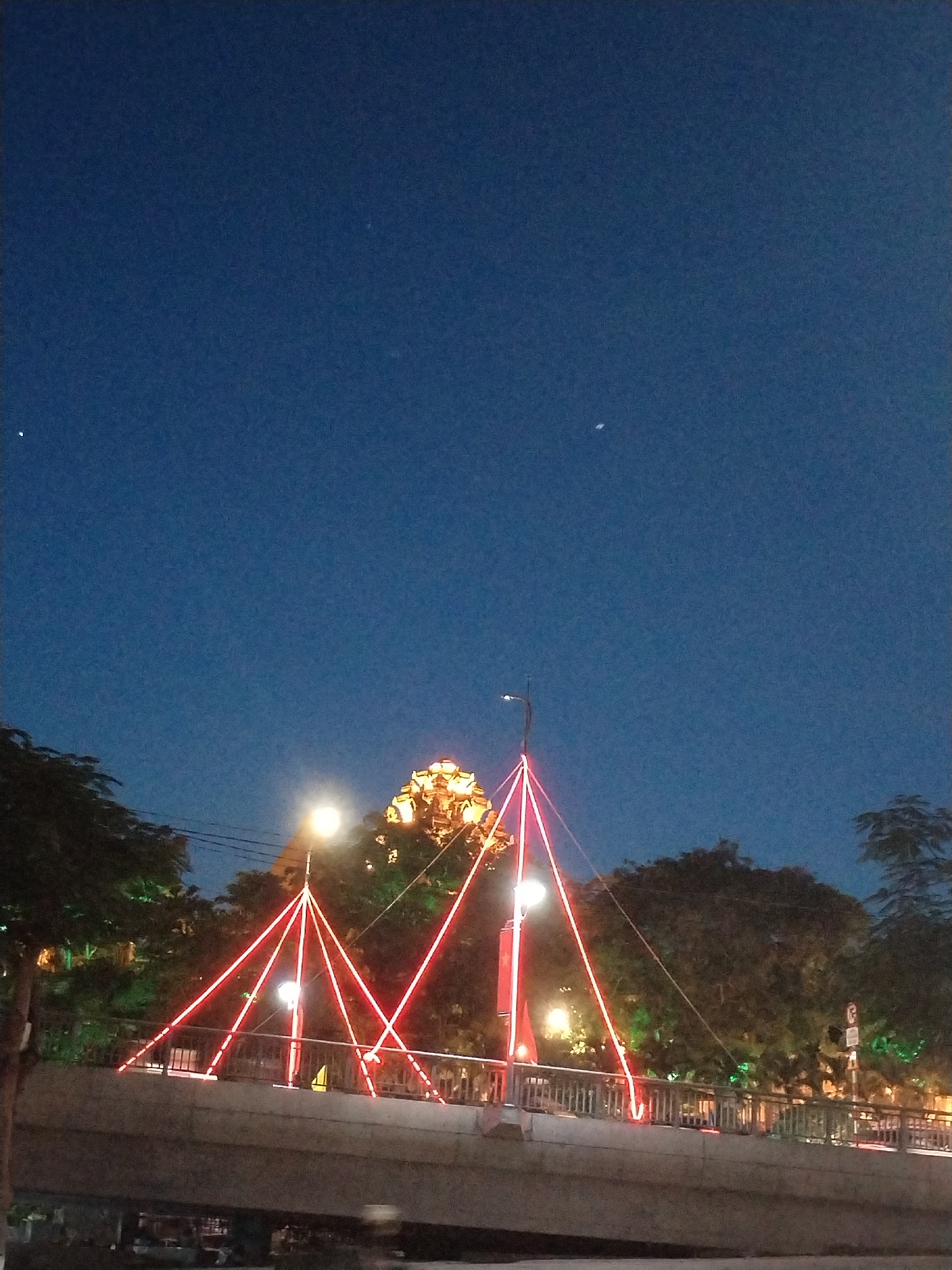 Captured at night in Bac Nha Trang Ward, Vietnam, the image features a bridge adorned with illuminated red triangle-shaped lights. The sky is a dark blue hue, punctuated by a few visible stars. Below, the bridge structure is visible, suggesting a lower vantage point. To the sides, trees line the scene, some with their own lighting, creating an interplay of light and shadow. A building with decorative lighting is visible behind the trees.