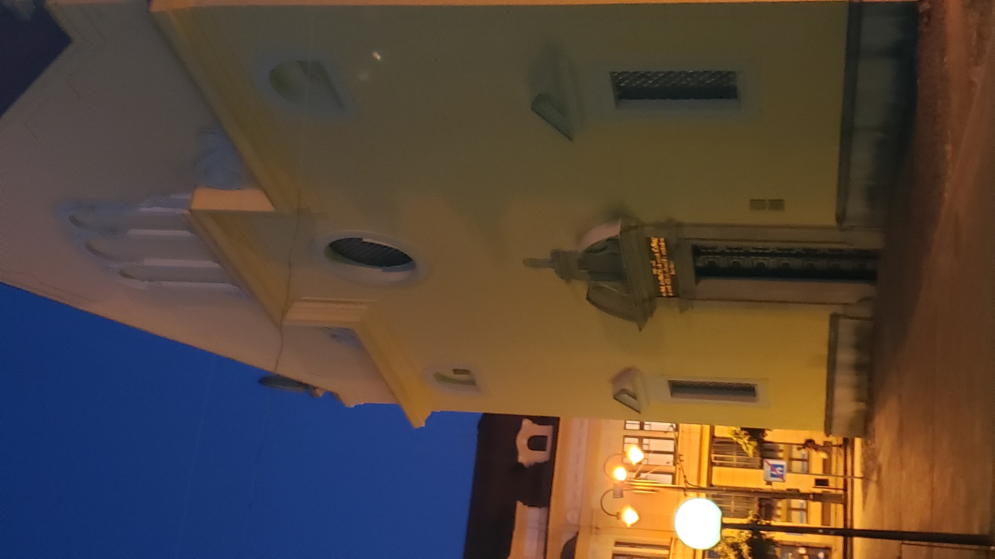 The image shows a historical building in the city of Prešov, Slovakia, likely a church, at dusk. The building's facade is painted a pale yellow and features arched windows and decorative architectural elements. A prominent entrance with a dark, ornate door is visible. To the left, another building with illuminated windows and streetlights is partially visible. The sky is a deep blue, indicating the transition from day to night. The scene appears to be a quiet urban environment.