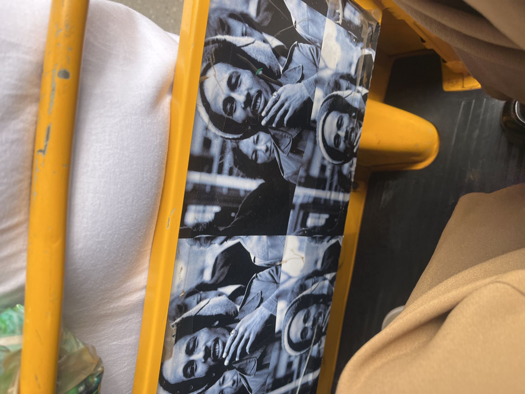 The image presents a close-up, tilted view of a yellow metal structure, possibly a support beam, railing, or part of a vehicle. Adorning a section of this yellow surface is a repeated pattern of a black and white photograph featuring the musician Bob Marley.
Bob Marley is depicted smiling, with his distinctive dreadlocks partially covered by a light-colored hat or beanie. He appears to be wearing a collared jacket, possibly denim, and a ring is visible on his left hand. Another individual, possibly a child, is partially visible behind him within the photograph. The repeated images show signs of wear, including scratches and creases, indicating they might be stickers or a decal that has seen use.
The yellow metal structure itself is utilitarian, exhibiting visible scuffs, chips in the paint, and general wear and tear. On the left side of the frame, a white, textured fabric is visible, likely clothing or a cushion. On the right, a light brown or beige fabric, possibly another piece of clothing or upholstery, is present. A dark, possibly black, surface is visible on the lower right, suggesting a floor or ground beneath the yellow structure. A green plastic bag is also faintly visible on the far left.
The scene captures a detail from an everyday environment, set in Galadimawa Village, Nigeria. There is no specific activity taking place beyond the subject of the photograph being displayed on the yellow structure. The lighting suggests it is daytime. No discernible text is visible within the image other than the implied subject of the photograph. The overall impression is of a casual, lived-in setting.