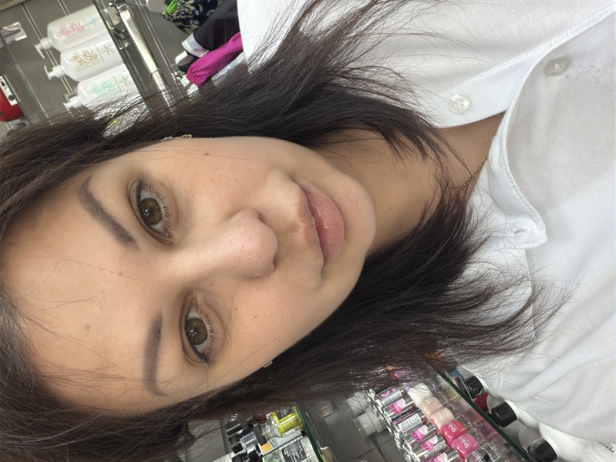 A head-and-shoulders selfie captures an adult female with long, dark brown hair and hazel eyes, looking directly at the camera. Her eyebrows are well-defined, and her full lips have a natural pink hue with a glossy finish. Her fair skin shows a few subtle freckles on her cheeks and forehead. She wears a white collared shirt, unbuttoned at the top to reveal a thin gold chain necklace. A small, clear stone earring is visible in her right earlobe.
The background, largely out of focus, indicates an indoor retail setting. On the left side, multiple shelves display various products, including small bottles with prominent "Alvin D'or" branding, typically associated with Russian cosmetics. On the upper right, several white pump bottles with distinct green, gold, and pink labels, featuring design elements like leaves and floral patterns, are visible. Some labels clearly state "Aloe" and "Olive." Below these bottles, a clothes rack holds various garments, including a bright pink item. The scene depicts a customer or employee within a beauty or general merchandise store, located in a retail store in the city of Sochi, Russia.