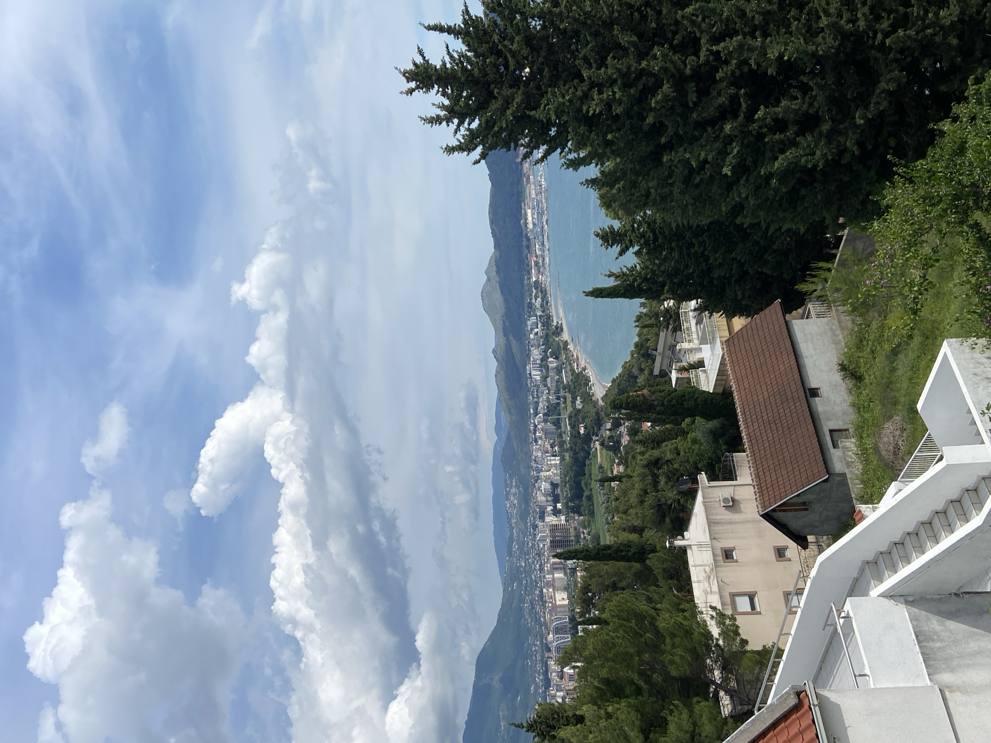 An eye-level, horizontal, exterior shot depicts a seaside view in Šušanj, Montenegro. The image, slightly canted to the left, prominently features the coastline stretching along the Adriatic Sea.
To the left, fluffy cumulus clouds fill a blue sky. On the right, mature trees frame the scene, partially obscuring residential buildings with brown and white exteriors. A white staircase, complete with a railing, descends in the foreground.
The city itself, a mix of residential and commercial structures, extends along the coast. Mountains rise in the distance, receding into the background. The sea appears calm, and the overall atmosphere suggests a sunny day.