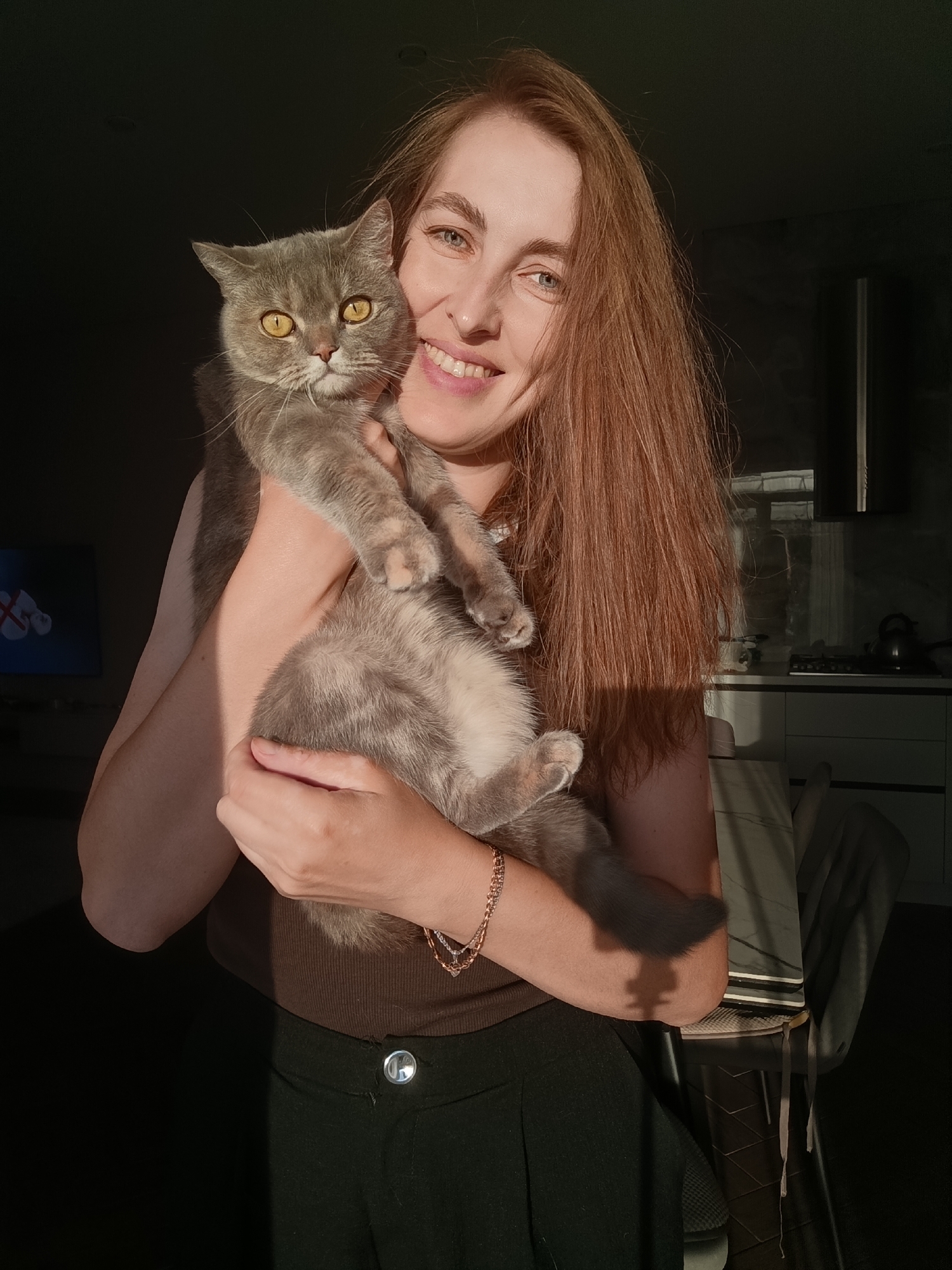 In this medium shot, a woman with long, reddish-brown hair smiles at the camera, holding a grey cat. The woman is wearing a brown ribbed top and dark pants with a silver button. A delicate gold bracelet adorns her left wrist. The cat, with bright yellow eyes, is cradled in her arms, its grey fur catching the sunlight.
The scene appears to be indoors, possibly a kitchen or dining area, suggested by the background which includes cabinets and a kettle on a countertop. The lighting indicates it is daytime, with strong sunlight casting dramatic shadows across the subjects and the room. The mood is warm and affectionate, conveyed by the woman's broad smile and the way she holds her pet. The location is Chaikovsky, Russia. There is no visible text in the image.