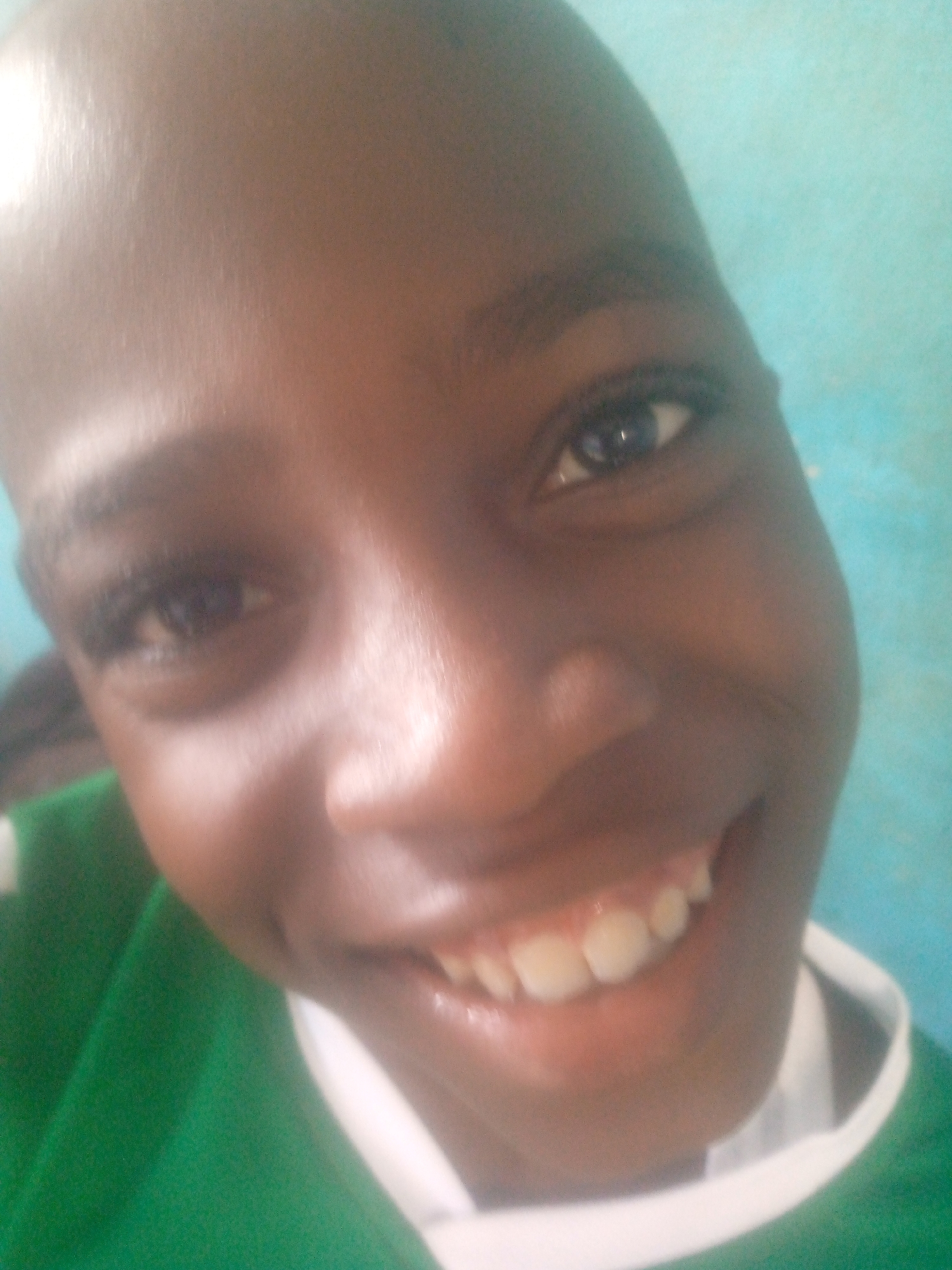 A close-up shot focuses on a dark-skinned person with a shaved head, capturing their smiling face. They are wearing a green shirt with a white collar. The location is unknown.