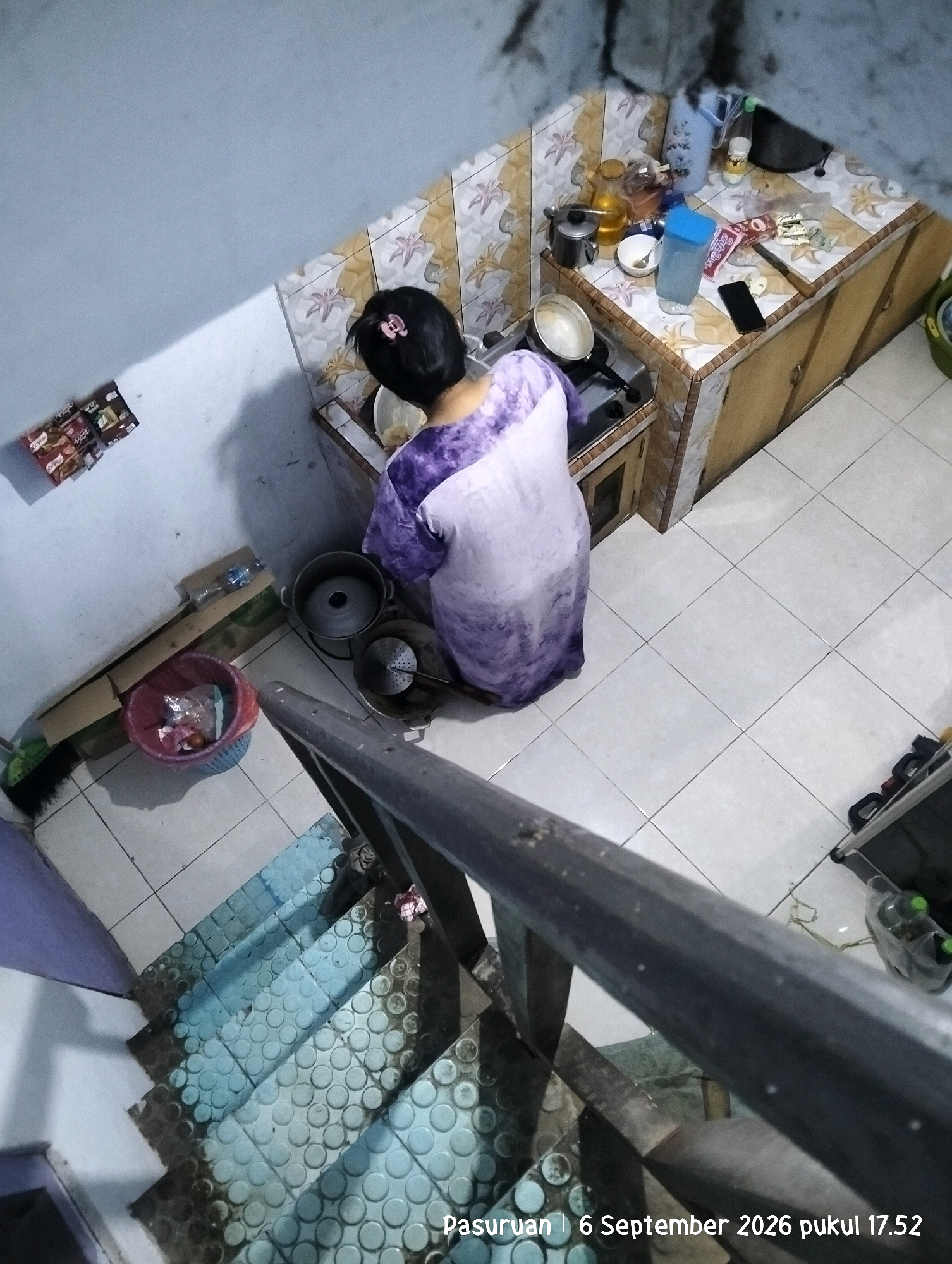 A medium shot captures an overhead view of a kitchen area within a home in Warung Dowo, Indonesia. A woman wearing a purple and white tie-dye smock is standing at a gas stove, preparing food. The kitchen is small and appears to be utilitarian, with tiled walls and countertops. Various kitchen utensils, pots, and containers are visible on the stove and countertop. To the left of the stove, a blue plastic trash can is overflowing with refuse. A set of stairs with blue-treaded risers is in the foreground, partially obscuring the kitchen scene. The overall lighting suggests it is either early morning or late afternoon. A timestamp in the lower right corner indicates the image was taken on September 6, 2026, at 5:52 PM.