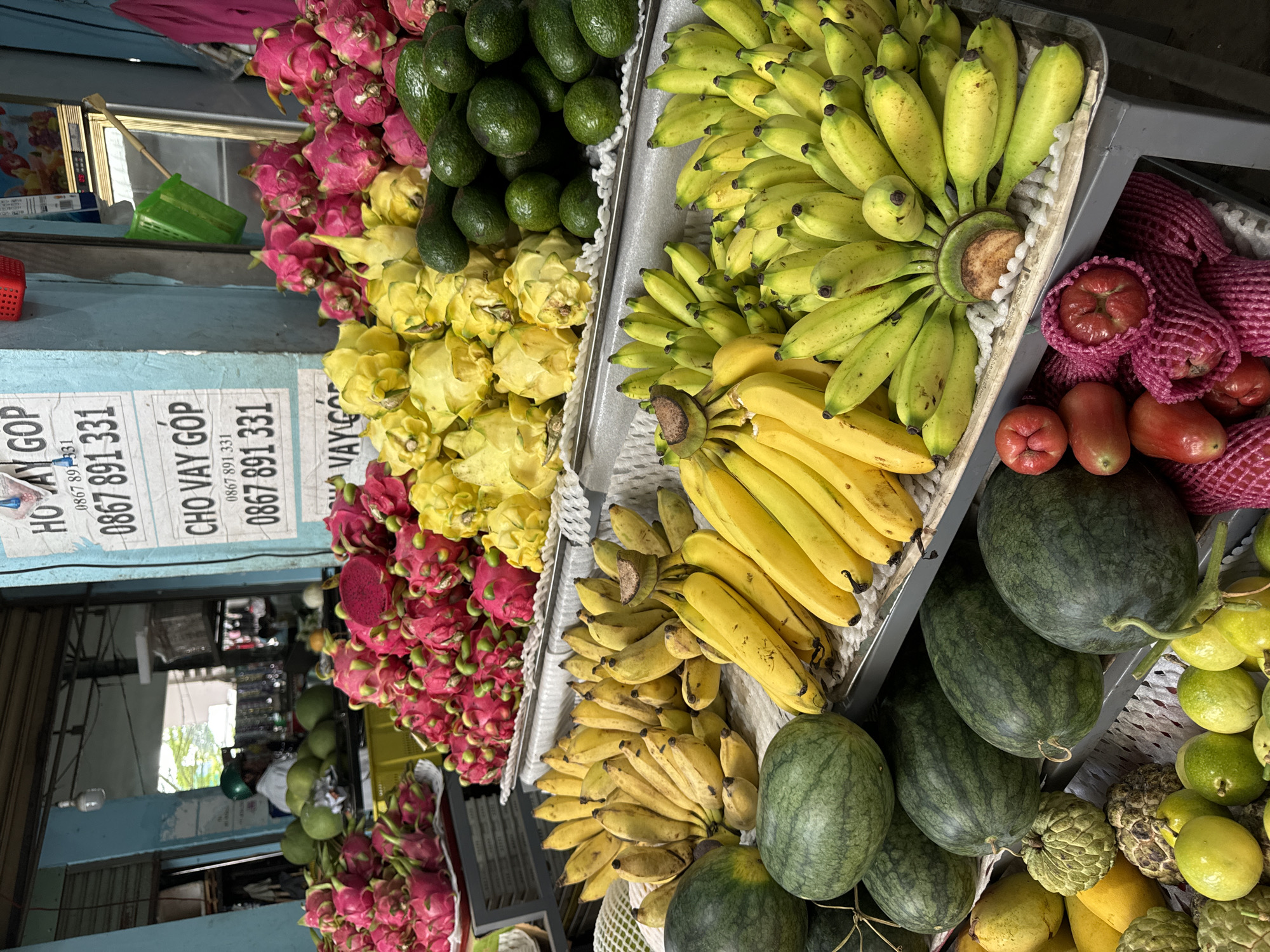A vibrant fruit market scene unfolds in Mũi Né, Vietnam. The foreground is dominated by a diverse array of fresh produce arranged on metal shelves. Prominently featured are numerous bunches of ripe yellow bananas, some with a slight green hue, and several large, dark green watermelons with distinctive lighter green stripes. Interspersed are green apples, red and pink dragon fruits, yellow dragon fruits, and green avocados. In the lower portion of the image, additional fruits are visible, including what appear to be custard apples and passion fruits, alongside some red, pear-shaped fruits. Behind the fruit displays, a stall with blue walls displays signage with Vietnamese text and phone numbers. A person wearing a green helmet is partially visible in the background, and the overall environment suggests a bustling outdoor or semi-outdoor market setting.