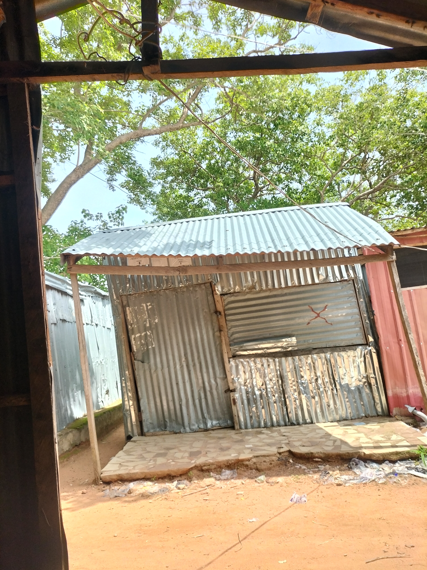 The image shows an outdoor scene, likely a makeshift structure or stall, in Jalingo, Nigeria. The structure has a corrugated metal roof supported by wooden beams and sides constructed from corrugated metal sheets. There is an opening in the front, revealing a rough, unfinished interior. A patch of red paint in the shape of an "X" is visible on a section of the metal wall. The ground in front of the structure is reddish-brown earth with scattered debris, including plastic bags. Behind the structure, lush green trees and blue sky are visible. To the left, another corrugated metal structure can be seen. The overall impression is of a simple, functional, and somewhat dilapidated construction.