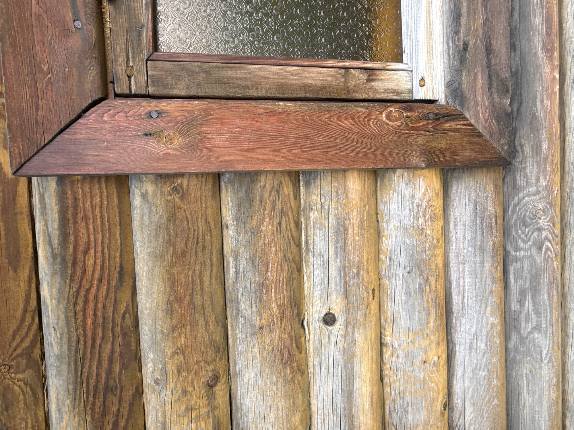 This is a close-up, eye-level, outdoor, daytime photograph of the corner of a wooden log cabin. The logs are horizontal and of varying widths and colors, ranging from light grey and weathered to rich brown with visible grain patterns and knots. A wooden window frame with a reddish-brown stain is visible on the right side of the image, angled across the logs. The window itself is partially visible and appears to have textured, patterned glass. There are visible nails securing the wooden elements. The image focuses on the textural details of the weathered wood. No people are present. The location is not specified in the image itself, but it is stated to be in Baryshevo, Russia.
