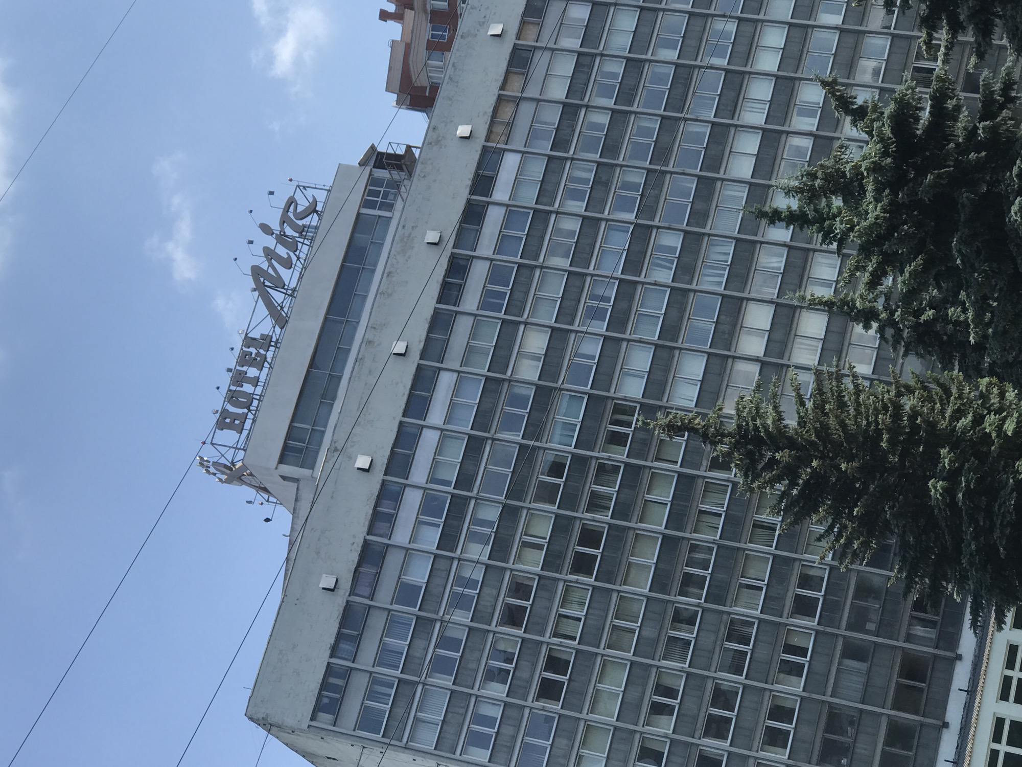 An outdoor, eye-level, medium shot shows a tall, grey, brutalist-style hotel building under a clear blue sky with a few wispy clouds. The building is composed of repeating rectangular windows and horizontal concrete bands. The hotel name, "HOTEL MIR," is visible in large, silver, serif font letters atop the building. Several dark green evergreen trees stand in the foreground, obscuring the lower portion of the hotel. Power lines are visible crisscrossing the sky.