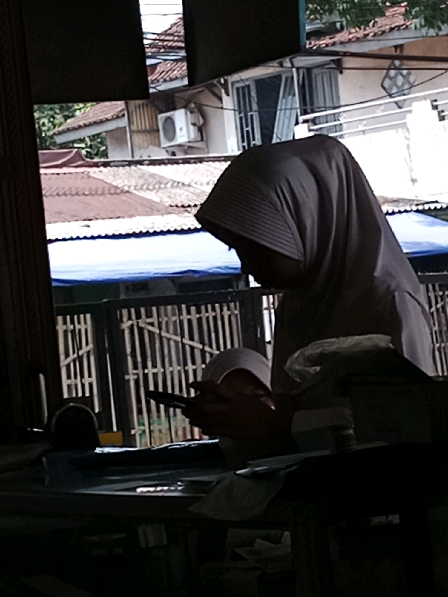 A side-view, medium shot captures a person wearing a grey hijab, looking down at a dark object, possibly a smartphone, held in their hands. The person is positioned in the foreground, their head and upper body silhouetted against a bright, outdoor background. Behind them, a white fence with vertical slats is visible. Beyond the fence, a blue tarp stretches across a surface, and further back, buildings with red tile roofs and white walls are seen. An air conditioning unit is mounted on one of the white walls. The overall lighting suggests an outdoor environment, possibly in the city of Muka, Indonesia, with a focus on the subject's engagement with the object in their hands.