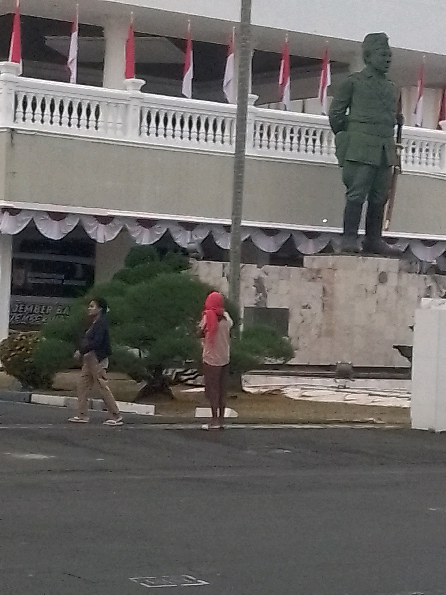 The image shows a street scene in front of a building adorned with Indonesian flags. In the background, a large statue of a man in military uniform stands prominently. In the middle ground, two individuals are walking across the street. The person on the left is wearing a dark jacket and light-colored pants, while the person on the right is wearing a light-colored shirt and a red headscarf. A green tree is situated between the individuals and the building. The building itself appears to be an official structure, possibly a government or administrative building, with a balcony and decorative railings. Text on the building appears to read "JEMBER," indicating the location as Jember, Indonesia. The ground in the foreground is asphalt, and there are white lines painted on it.