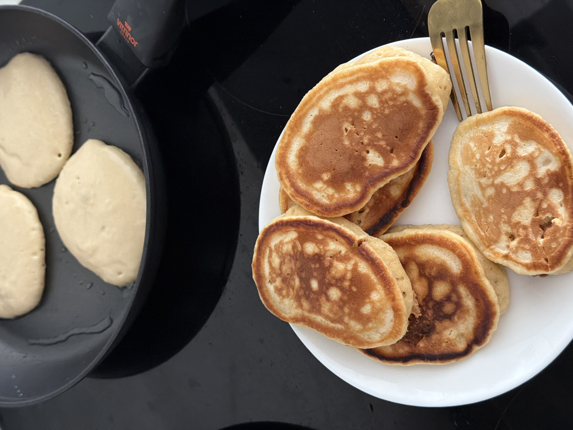 In an indoor setting, likely a kitchen, on a black stovetop, a non-stick frying pan contains at least two uncooked pancakes. The pancakes are round and pale yellow, with a slightly viscous liquid, possibly oil or butter, pooling around them. The frying pan has a black handle with the red logo "VITRINOR" visible. In the foreground, a white plate holds a stack of at least five golden-brown, cooked pancakes. The pancakes are irregular in shape and have a slightly bumpy texture. A single gold-colored fork rests on the edge of the plate, touching one of the pancakes. The background is dark and reflective, suggesting a modern kitchen surface. The scene does not provide any information about the city of Finestrat, Spain.