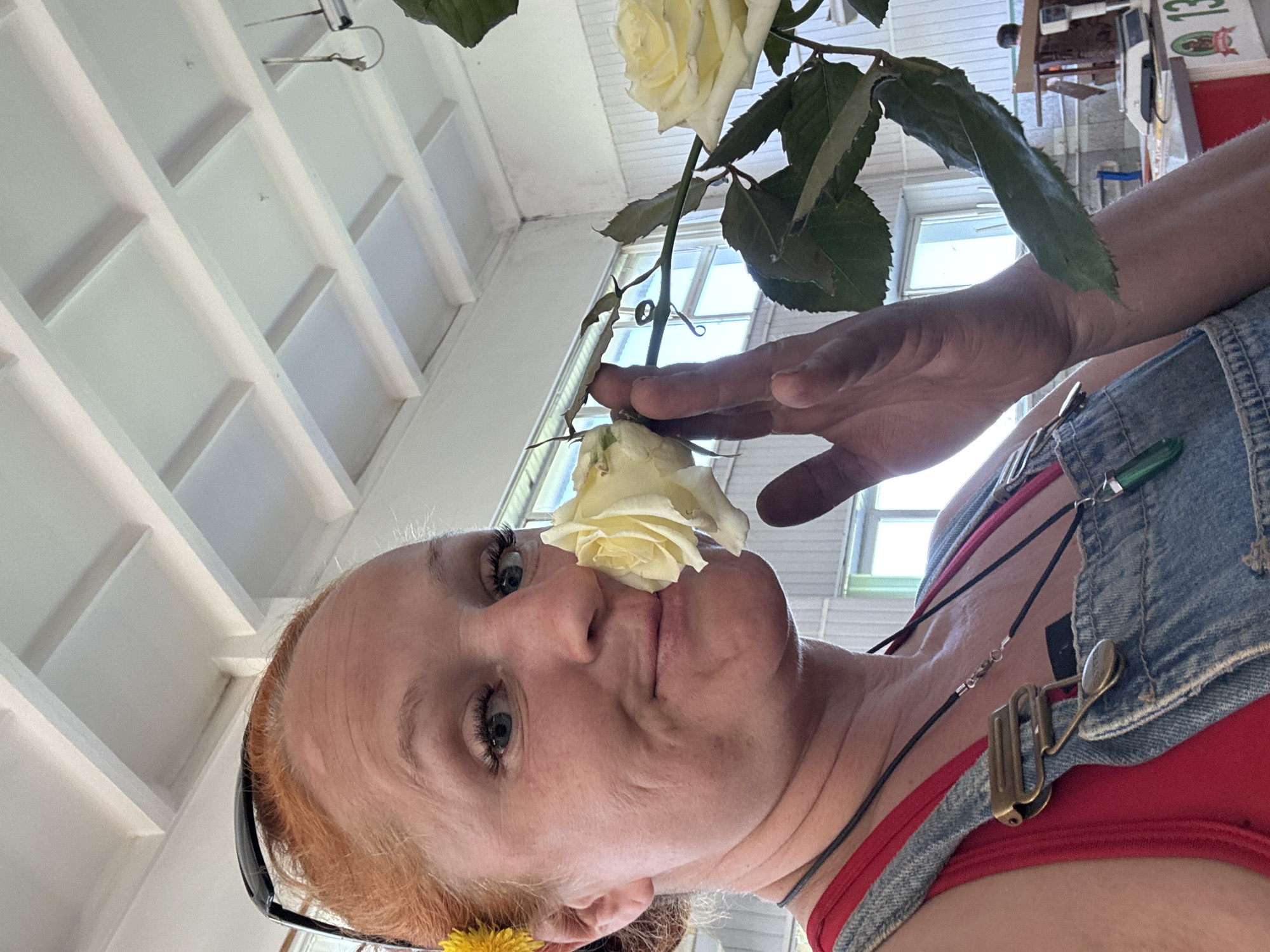 A woman with red hair poses with a white rose touching her nose. She is wearing a red tank top and denim overalls with metal buckles. A pair of sunglasses are tucked in her hair. The background consists of a white latticed ceiling and windows. It is difficult to confirm the location as Chaikovsky, Russia from the image alone.
