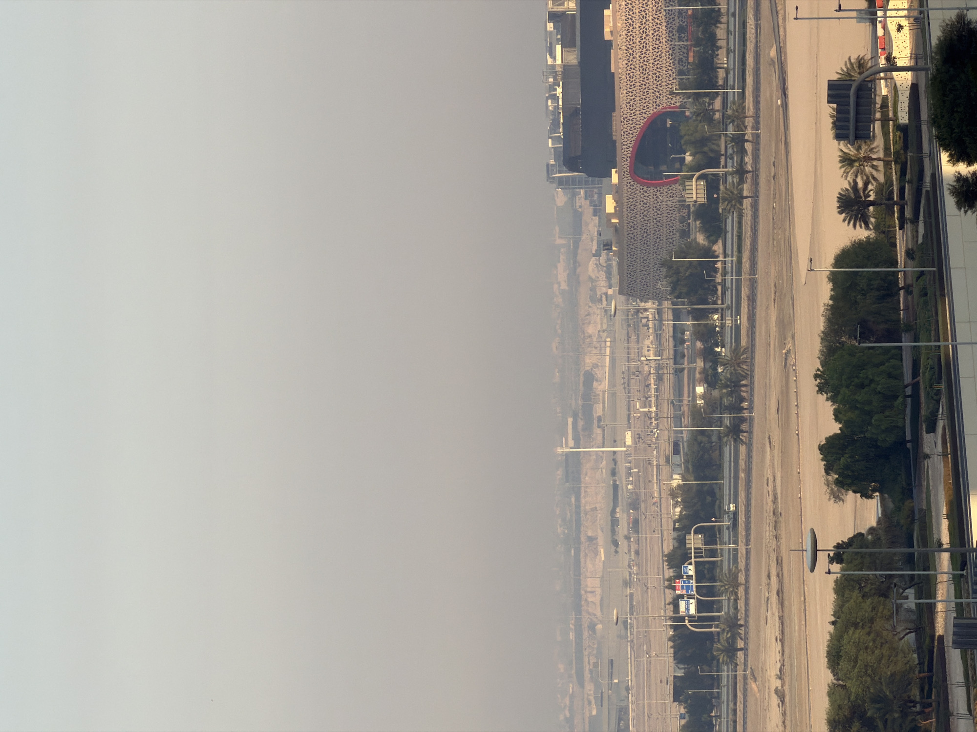 A high-angle, wide shot captures an urban landscape under a hazy, light blue sky. In the foreground, a dark-colored building with white trim runs along the bottom edge of the frame. Beyond that, a network of roads and streetlights is visible, with patches of green trees and palm trees interspersed. Several street lamps, some with modern, circular tops, dot the scene. In the midground, a large, modern building with an ornate, patterned facade and a prominent red, arched opening stands out. This building is surrounded by flat, sandy terrain and more sparse vegetation. Further in the distance, a vast expanse of the city stretches out, characterized by low-rise buildings and industrial structures, all softened by atmospheric haze. A tall, slender white tower is visible in the far distance. The overall impression is one of a sprawling, modern city on a day with reduced visibility.
