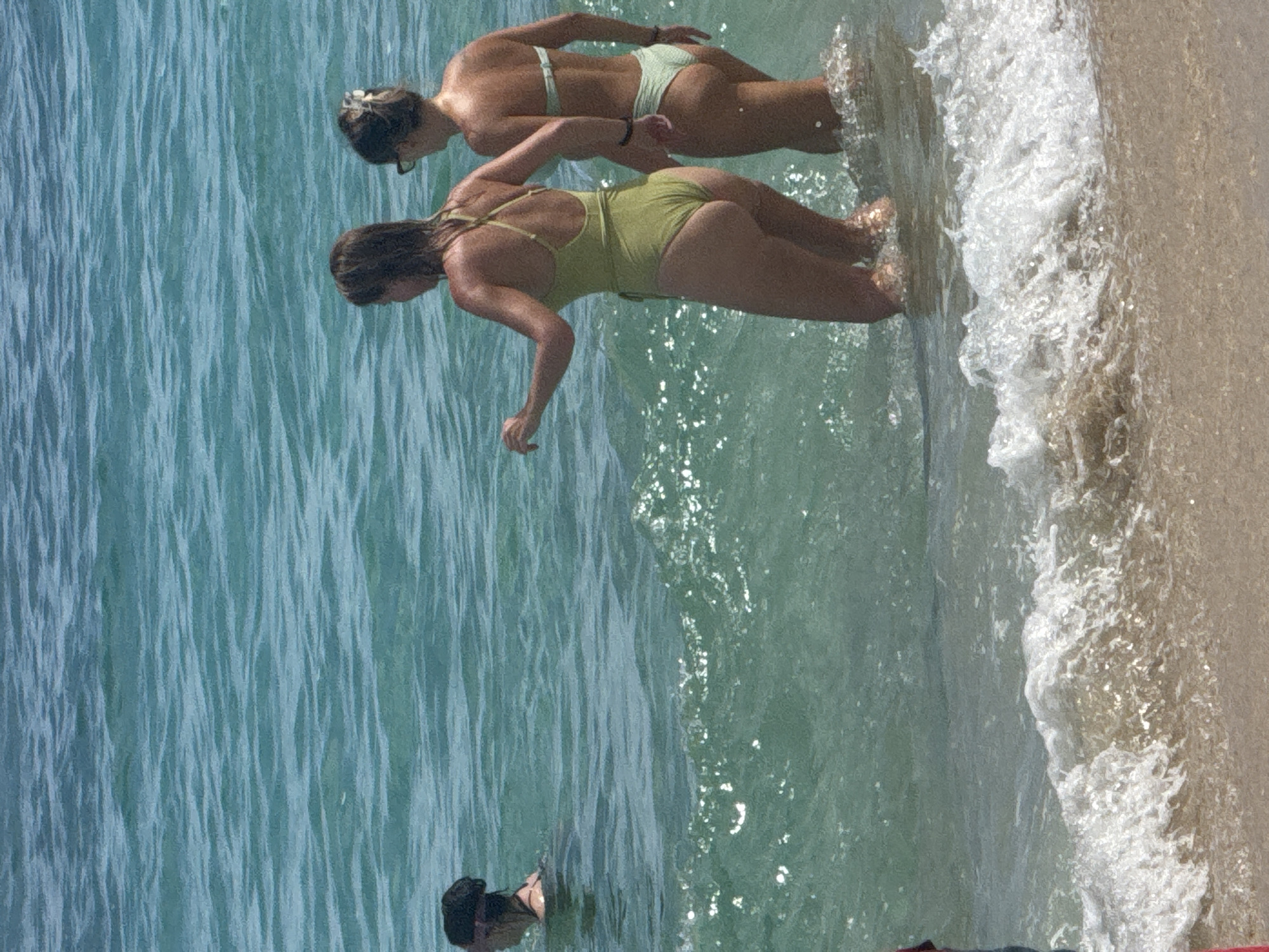 Two women in swimwear stand waist-deep in the ocean along a sandy beach. The woman closer to the camera wears a one-piece swimsuit, while the other wears a two-piece bikini. Waves are visible breaking on the shore. The water is clear and turquoise. The photo was taken in Kuta Selatan, Indonesia.