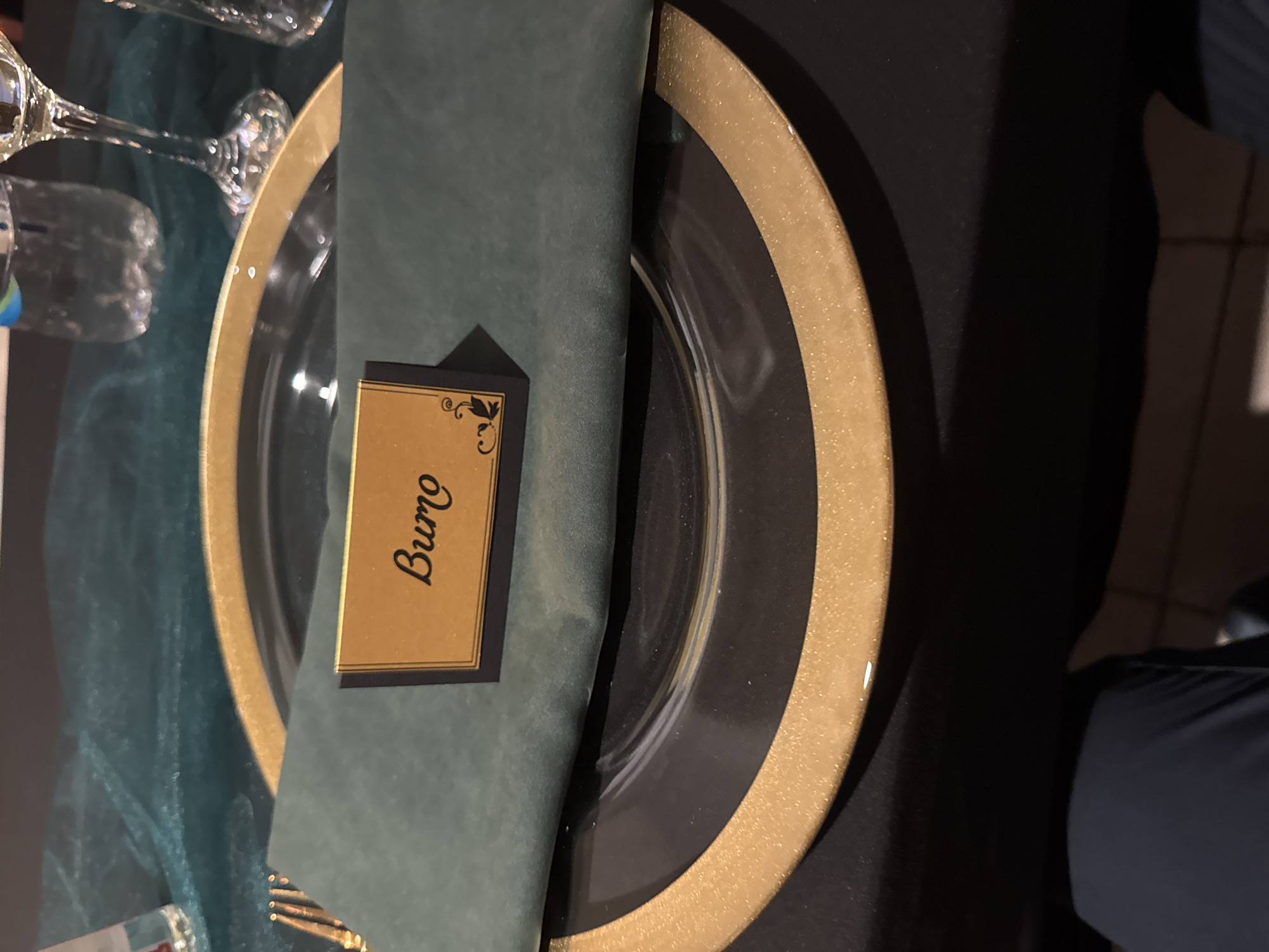 The image shows a place setting, likely at a formal event. There is a clear glass charger plate with a gold rim. A folded dark green napkin is placed on the plate. A name card with gold lettering, displaying the name "Buro," rests on top of the napkin. A wine glass is visible in the upper left corner of the image. The tablecloth appears to be dark teal. The presence of the formal setting implies that this image could be associated with an event like a wedding or formal dinner that might have occurred in Thermi, Greece. A person wearing navy blue pants is visible on the right side of the frame.