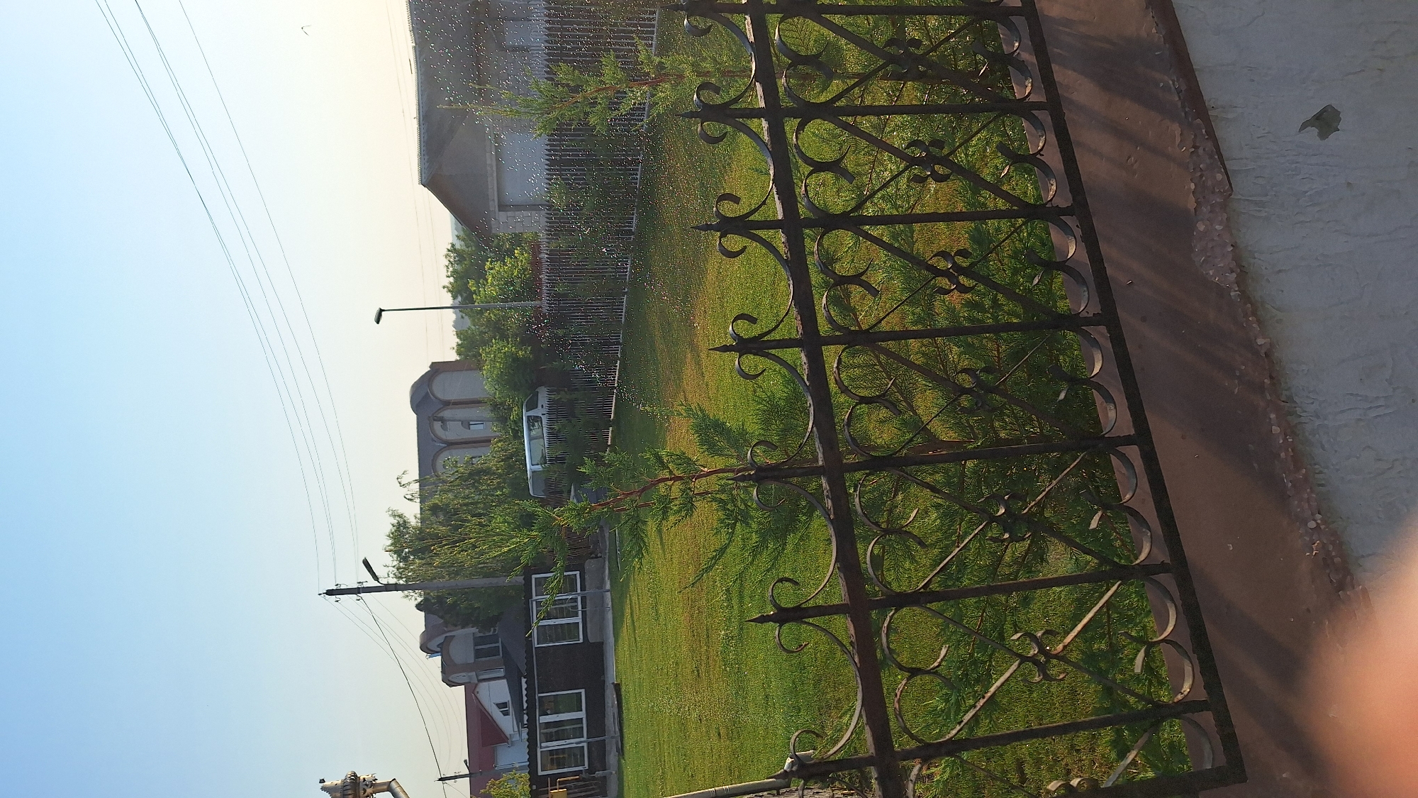The image shows a residential property in Sărata-Galbenă, Moldova, on a clear, sunny day. In the foreground, a black wrought-iron fence runs horizontally across the bottom of the frame, partially obscuring a lush green lawn. Two small evergreen trees are visible behind the fence, growing on the lawn.
Beyond the lawn, a dark-colored building with large windows is situated on the left side of the property, likely a house or a garage. Further back, a white van is parked. A tall fence, also dark in color, runs along the back of the property, separating it from neighboring areas.
In the background, several houses with distinctive arched rooflines and brickwork are visible, suggesting a typical village or suburban architectural style in the region. Electrical wires and utility poles are strung across the sky, indicating the presence of infrastructure. A streetlamp stands near the center of the background. The sky is clear blue, with a slight haze near the horizon, and the sun is casting shadows, indicating it is likely morning or late afternoon. The overall scene depicts a tranquil and orderly residential setting.