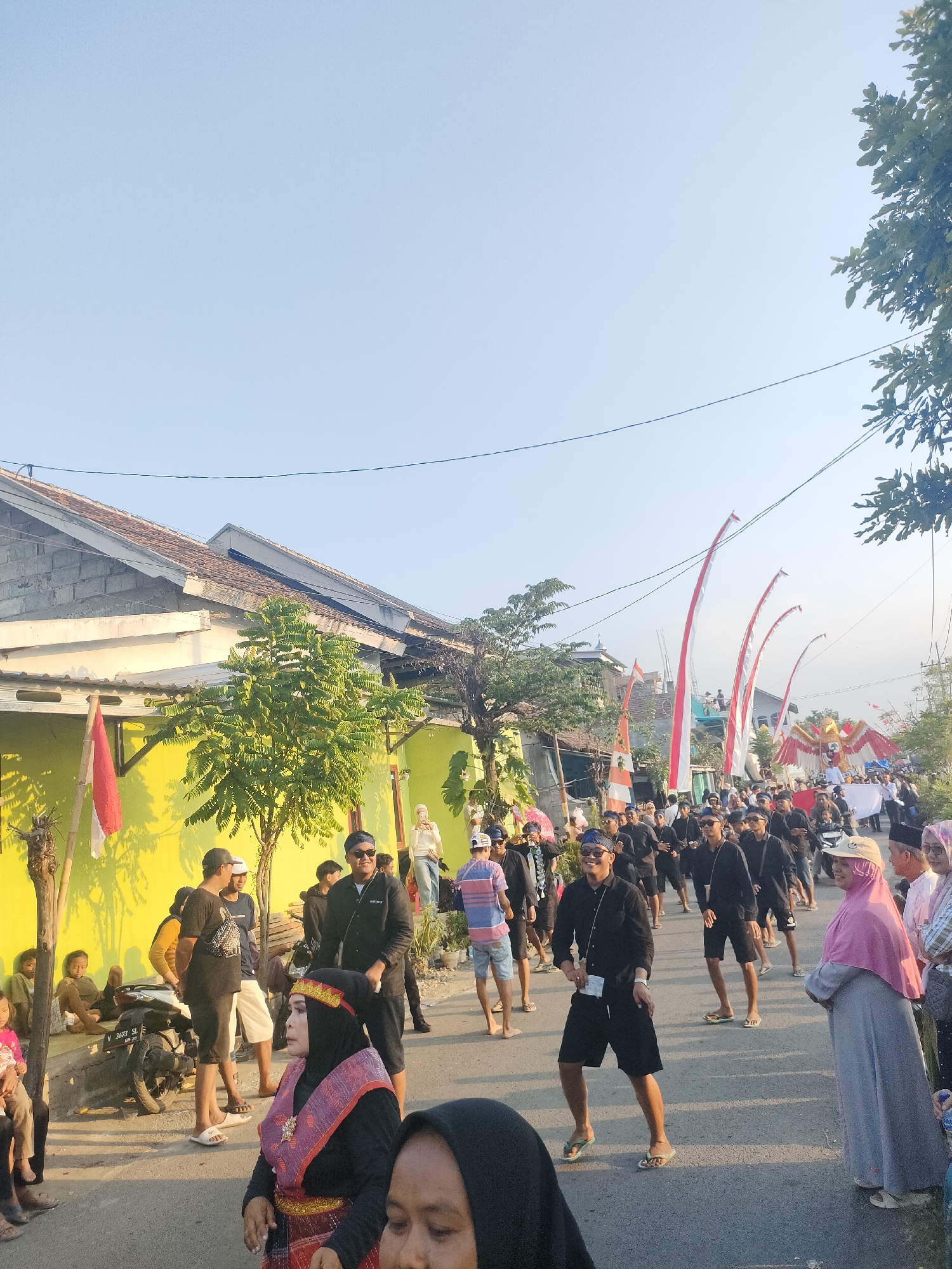 In Tongaswetan, Indonesia, a lively outdoor scene unfolds on a street lined with single-story buildings, their walls painted in bright yellow. The sky above is a clear, pale blue, with a few thin wires strung across. A group of people are gathered, many dressed in black attire, participating in what appears to be a procession or celebration. Tall, red and white striped banners with a pointed top are erected along the street, adding to the festive atmosphere.
In the foreground, to the left, a woman in traditional attire, featuring a black top with colorful patterned fabric around her shoulders and waist, and a black headscarf with a decorative band, is looking towards the right. Beside her, to the right, another woman in a light pink hijab is also visible. In the middle ground, a man in a black shirt and shorts, wearing sunglasses and a blue headband, is captured mid-movement, smiling. He is part of a larger group of men in similar black outfits, some walking, some appearing to dance.
Further back, on the left side of the street, a motorcycle is parked. A few individuals are seated and standing near the buildings, observing the event. The buildings have tiled roofs, and some have open windows. To the right, a large, colorful eagle-like effigy is being carried, adorned with red and white. The ground is paved, suggesting a residential or communal street. The overall impression is one of a vibrant cultural event taking place in a warm, sunny environment.