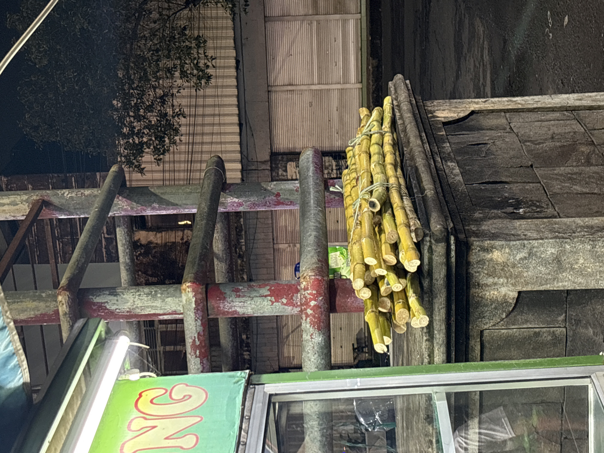 The image presents a nighttime street scene in Jember, Indonesia, centered around what appears to be a sugarcane juice vendor's stall. A prominent bundle of freshly cut, yellow-green sugarcane stalks, tied with twine, is displayed on the right, suggesting they are ready for sale or processing.
The sugarcane hangs near a sturdy, weathered structure composed of metal poles and horizontal beams, which exhibit chipped red and white paint and considerable rust. Below this structure, a portion of a green sign is visible, bearing red and yellow lettering that spells out "ONG," likely part of a larger business name. A bright, white fluorescent light fixture illuminates the area, confirming the late hour.
In the upper left background, a dark tree with dense foliage and visible aerial roots stands against the night sky. Behind the metal framework, a corrugated metal shutter or garage door can be seen, hinting at an adjacent building. To the right of the sugarcane, a dark, textured wall, possibly made of stone or rough concrete, forms the background. The bottom of the image reveals part of a glass display case or window belonging to the vendor's stall.
The scene depicts a typical roadside food setup, where fresh sugarcane juice is commonly sold. The absence of people indicates either an off-peak time or a temporary moment of inactivity. The overall impression is one of a functional, no-frills street vendor operation.