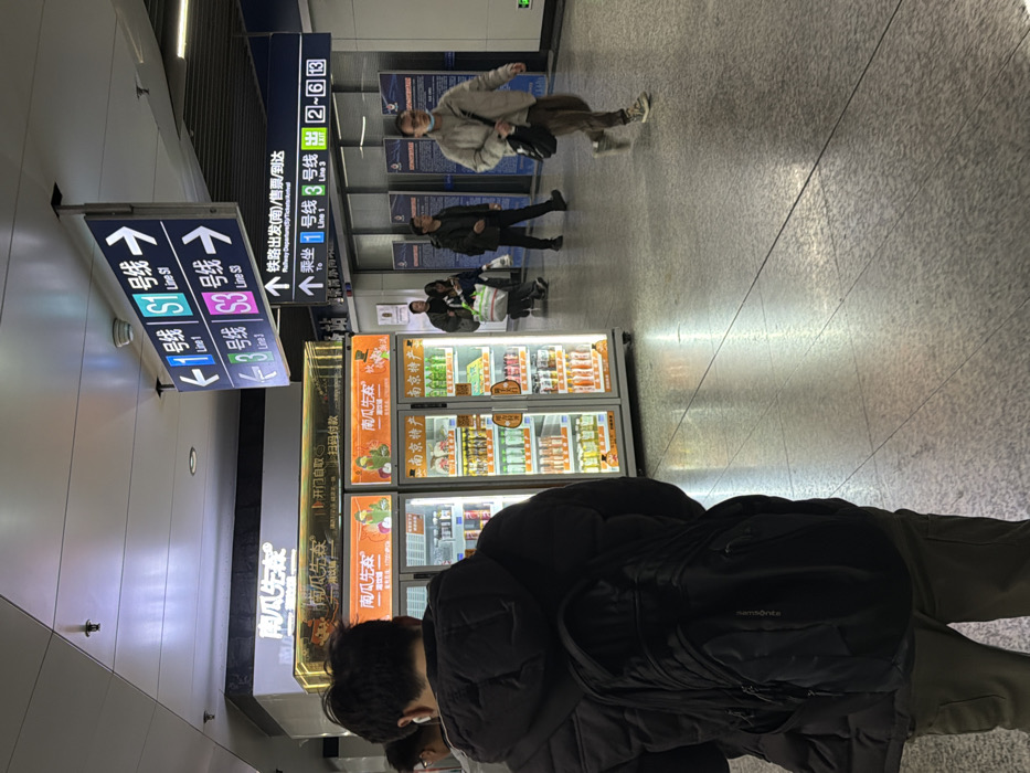 This image depicts an indoor public concourse within a metro station, identified as being in Yuhuatai District, Nanjing, China, by the presence of Nanjing Metro Line S1 and S3 signage and "Nanjing Specialty" branding. The environment features a polished, light-grey speckled floor and a white ceiling with recessed lighting.
In the left foreground, a male individual is observed from behind, wearing a black puffer jacket and olive-green trousers, with a black "sportporter" backpack. White wireless earbuds are visible in his ears. He stands facing two brightly lit, glass-fronted vending machines branded "南瓜先生" (Mr. Pumpkin), which display various bottled beverages and packaged items, including "南京特产" (Nanjing Specialty). An illuminated sign above the machines indicates "开门自取 / 扫码付款" (Open door and take / Scan code to pay).
In the mid-ground and background to the right, several individuals are present. One male subject walks from right to left, wearing a light-colored jacket, brown trousers, glasses, a light-blue face mask, and carrying a black shoulder bag. Other subjects are walking or standing further in the background. Overhead, a large blue and white directional sign points left to "1号线 Line 1" and "S1线 Line S1," and right to "3号线 Line 3" and "S3线 Line S3." Below this, another blue sign directs to "铁路出发(站)/售票/到达" (Railway Departure (Station)/Ticket/Arrival) and "乘车 1号线 3号线 出 2~6" (To Take Line 1 Line 3 Exit 2~6), indicating a connection to a railway station. The scene is evenly illuminated, casting faint reflections on the floor.