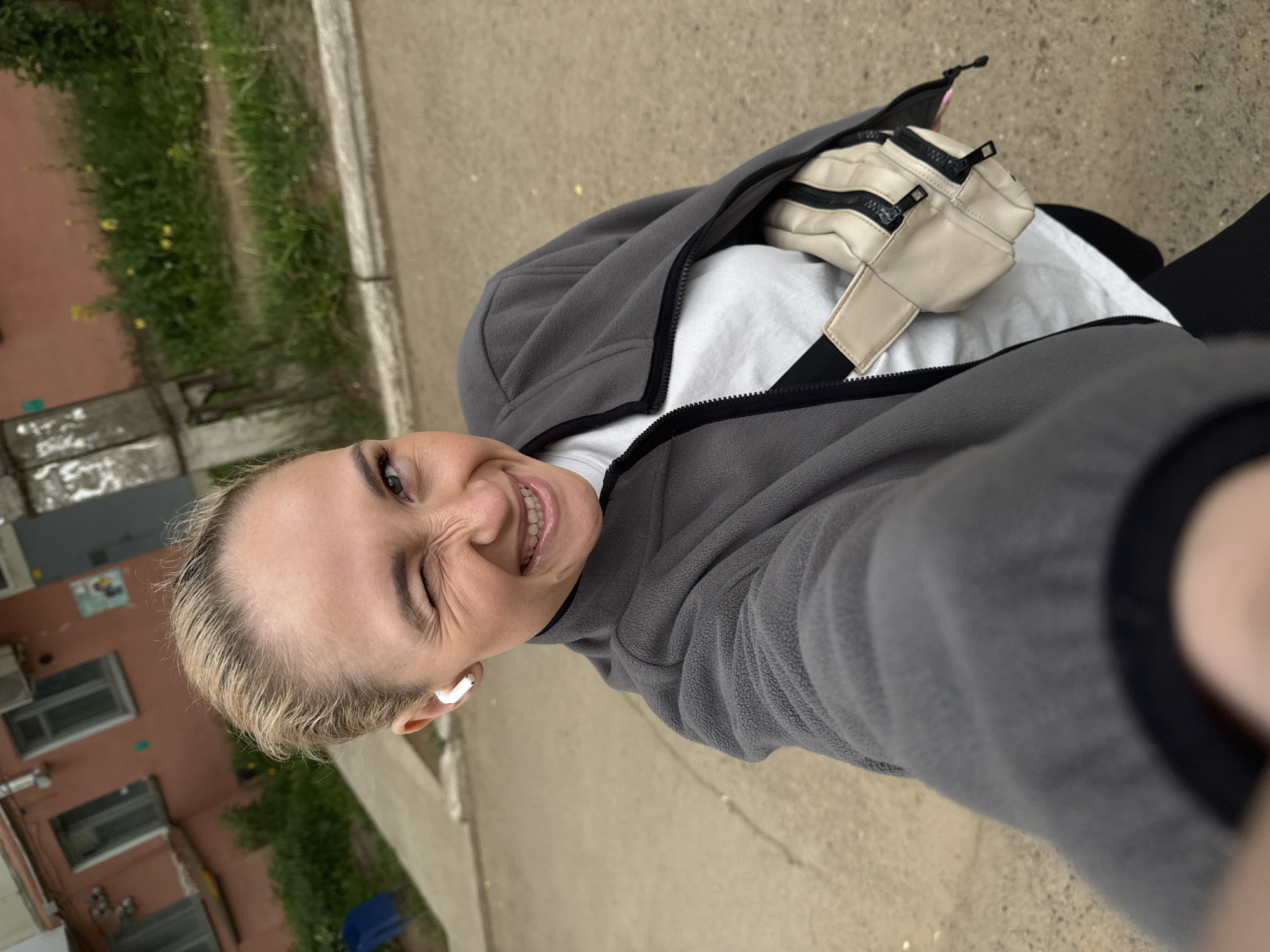 A cheerful woman is captured in a close-up, tilted selfie, looking down at the camera with a wide, bright smile and a wink. She has short, light blonde hair and is wearing a white earbud in her left ear. Her attire consists of a grey fleece zip-up jacket over a white t-shirt. A beige fanny pack with black zippers is worn across her chest, partially visible beneath the jacket.
The setting appears to be outdoors on a paved surface, likely a sidewalk or path. To the left, there is a strip of green grass and foliage, and further back, a multi-story building with a light reddish-orange exterior and windows. A concrete curb separates the paved area from the green space. The lighting suggests it is daytime, with diffused light indicating either an overcast sky or an indirect light source, as no harsh shadows are present. The woman's expression conveys happiness and playfulness. No visible text is present in the image. This scene is set in an urban or residential environment, with the provided context placing it in Chaikovsky, Russia.