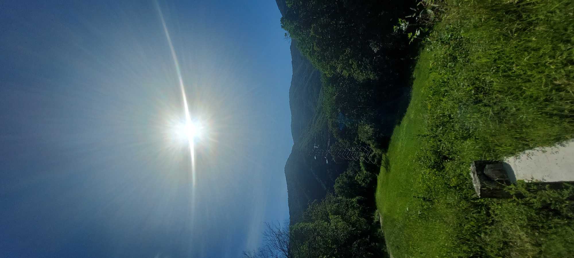 The image shows a landscape in Bachkovo, Bulgaria, featuring a bright sun in a blue sky, partially obscured by atmospheric haze. To the right, a mountain slope is covered in dense green vegetation and trees. There's a tall transmission tower in the middle of the mountains. In the lower right corner, a grassy area with a concrete structure is visible.