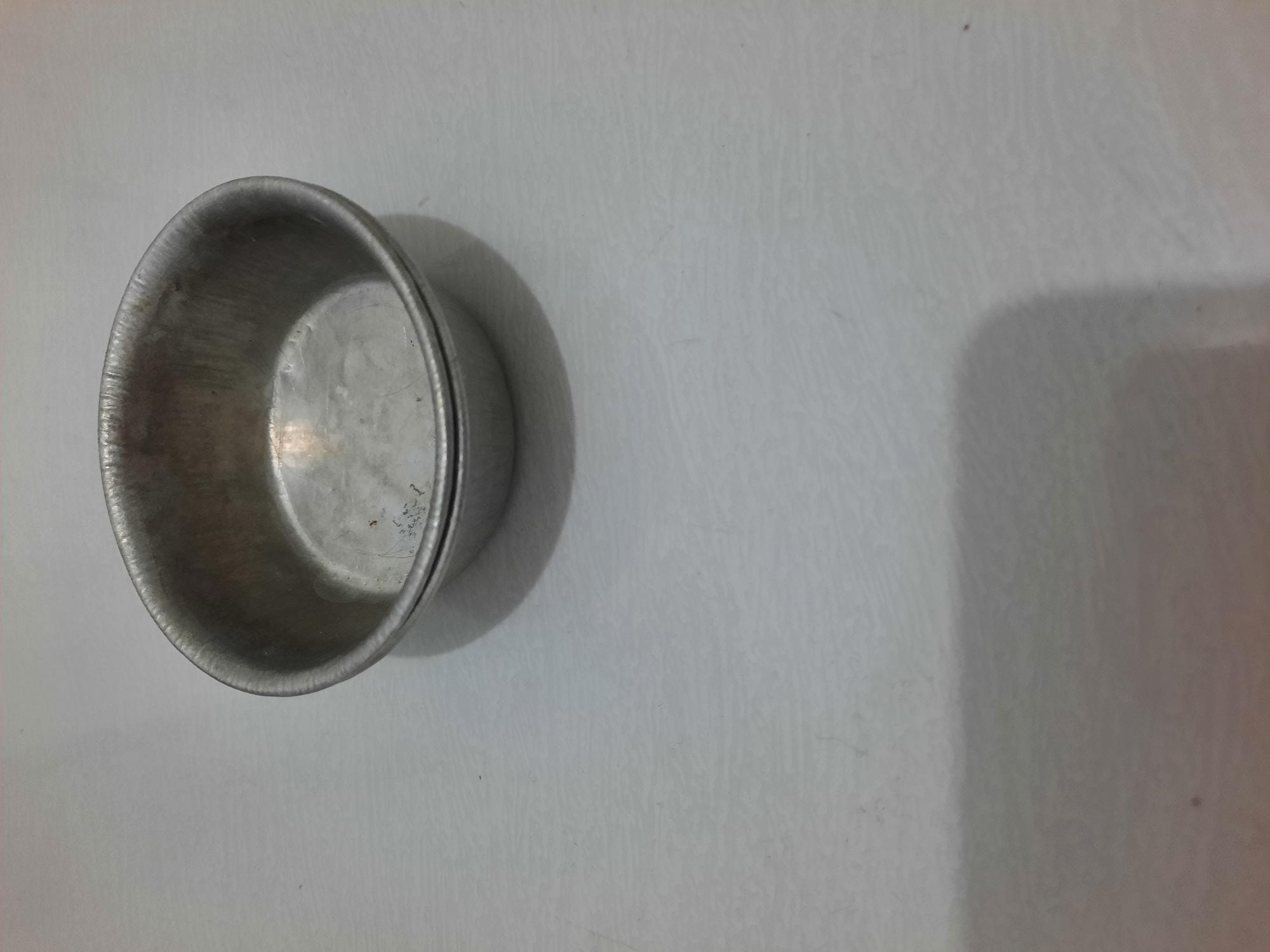 The image shows a single, empty, silver-colored metal bowl placed on a light-colored, textured surface. The bowl is positioned in the lower left quadrant of the frame, casting a shadow to its right. The surface appears to be a tabletop or countertop, with a subtle pattern of faint lines. The lighting suggests an indoor environment, possibly with natural light coming from the right, creating a soft shadow. There is no visible text in the image. The overall scene is simple and static, with no activity taking place.