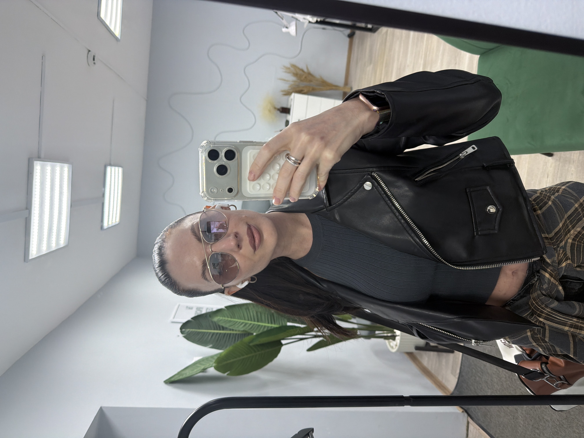 A woman takes a selfie in a room with a large houseplant, wearing a black leather jacket, gray ribbed top, sunglasses, and plaid shirt tied around her waist. She holds a smartphone with a textured case. A light fixture is visible in the background along with a wall with a decorative design. The location appears to be in an indoor space, potentially in the city of Perm, Russia.