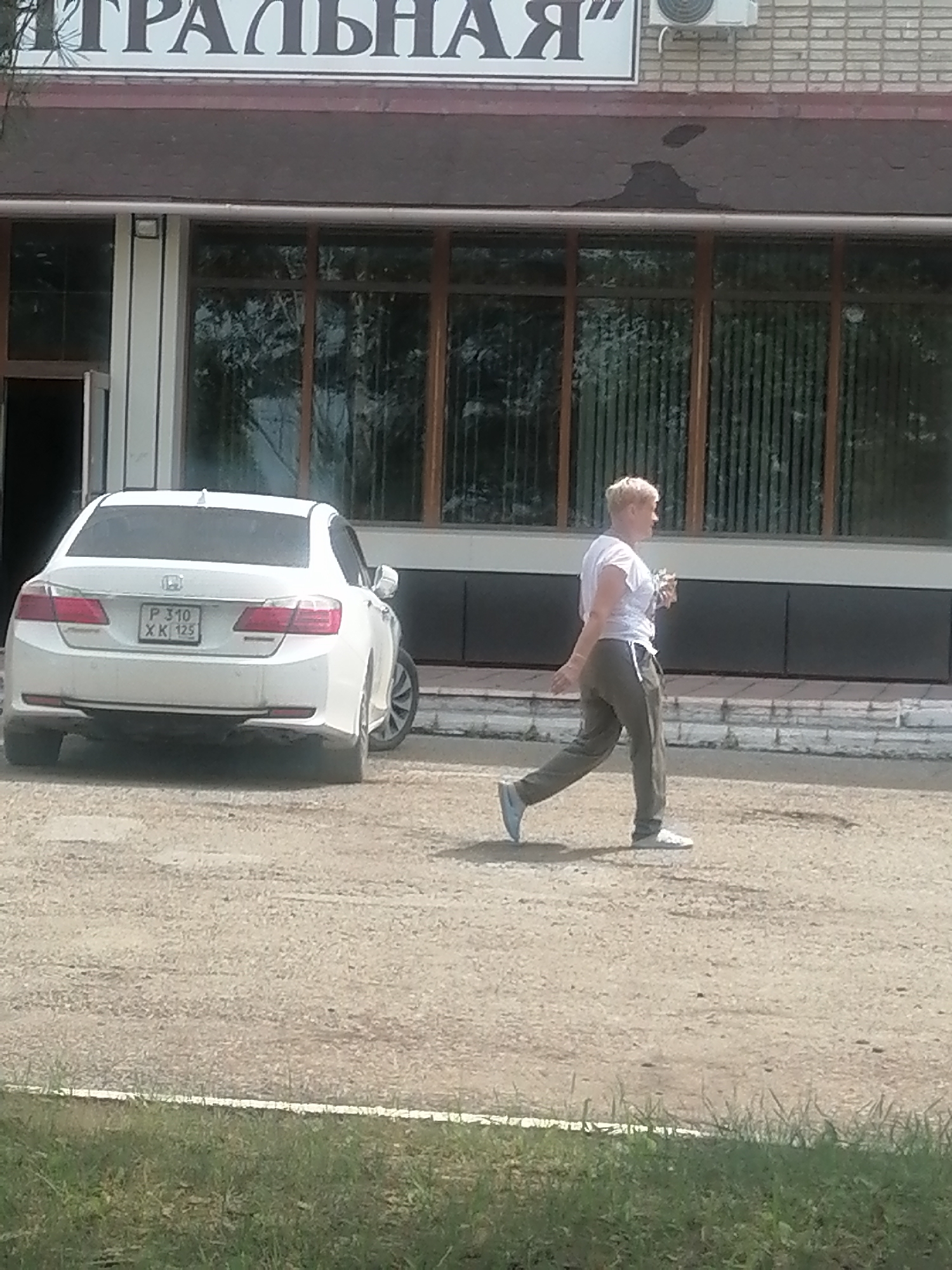 A blonde woman with short hair, wearing a white t-shirt and gray sweatpants, walks from left to right across a gravel lot. She is holding a small item in her right hand. Behind her, a white Honda Accord sedan is parked. The car has a license plate that reads "P 310 XK 125". In the background, a building with large windows and a sign with Russian text is visible. The sign appears to say "ТРАЛЬНАЯ". An air conditioning unit is mounted on the brick wall above the building's entrance. The scene appears to be in a commercial or public area. The ground is covered in gravel and there is a strip of green grass in the foreground. The weather is clear and sunny.