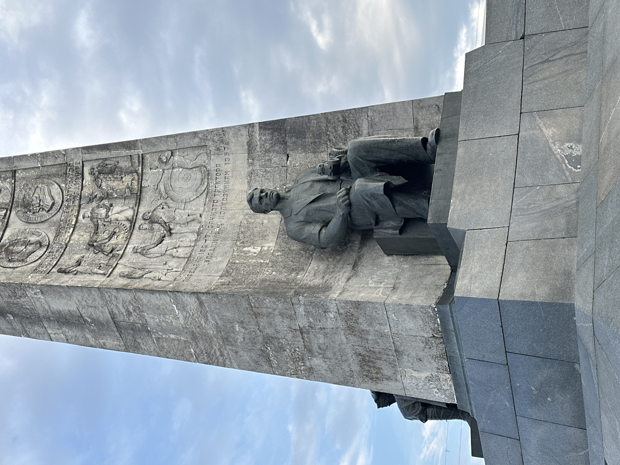 The image shows a monument in Vladimir, Russia. A bronze statue of a man sitting and holding a scroll is mounted on the side of a tall, gray stone obelisk. The obelisk is decorated with relief carvings depicting scenes of agricultural and industrial progress. The monument is situated against a cloudy sky. The right side of the monument consists of dark grey, smooth stone blocks.