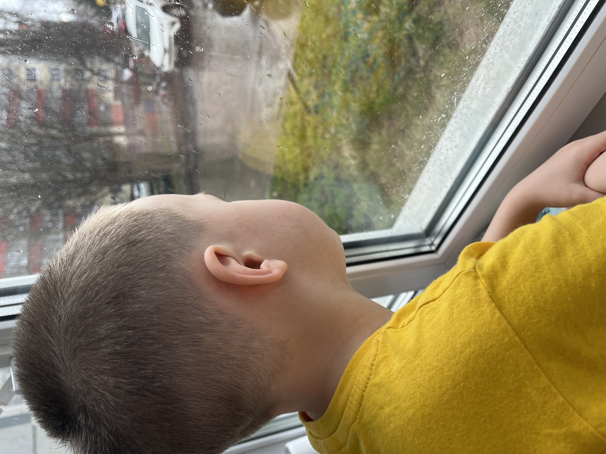 A young child with a buzz cut and wearing a yellow shirt is looking out a window with water droplets on the glass. The view outside the window is blurred, showing indistinct buildings and trees, reflecting a rainy or wet day. The child's face is tilted upward, with the ear and side of the head in clear view. The location cannot be confirmed as Usman, Russia, based on the visual information.