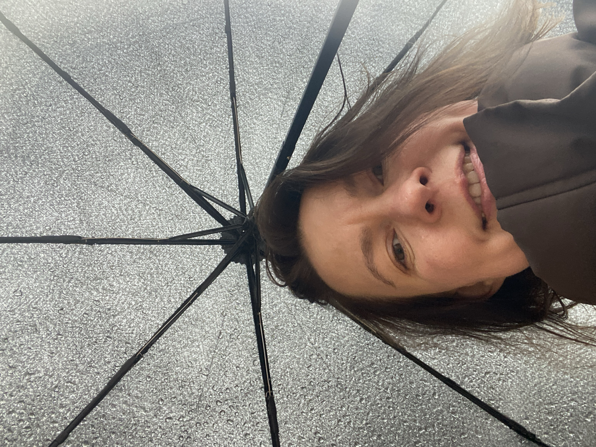 This close-up, low-angle shot captures a woman's face partially visible on the right side of the frame, looking up through a translucent umbrella. The umbrella's internal structure, with its dark radial spokes and a canopy covered in numerous small water droplets, fills most of the image, particularly the left and center. The woman has dark hair and a light complexion, displaying a wide, cheerful smile that shows her teeth. Her right eye is clearly visible, and she appears to be wearing a dark brown jacket or coat.
The water droplets on the umbrella indicate rainy weather, and the woman is likely using it for protection. The diffused lighting suggests an overcast daytime. Her expression conveys a sense of amusement or happiness. While the provided context places the image in Chaikovsky, Russia, there are no specific geographical cues within the image itself. There is no visible text.