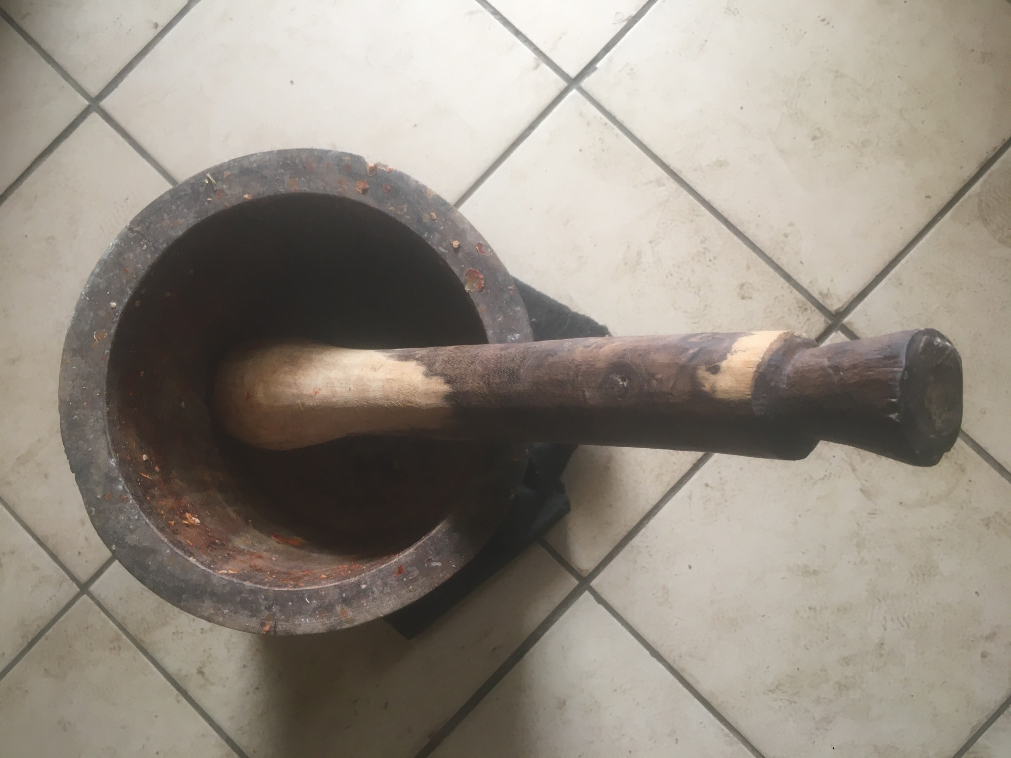 The image depicts a wooden mortar and pestle on a tiled floor. The mortar is a large, cylindrical vessel with a dark, weathered exterior and a visible reddish-brown interior. The pestle, used for grinding, is situated inside the mortar; it is primarily light-colored towards the grinding end and dark brown with a distinct handle on the other end. The tiles are a light color with dark grout lines. The object appears to be located indoors. There is no indication this is in Gwarinpa, Nigeria.