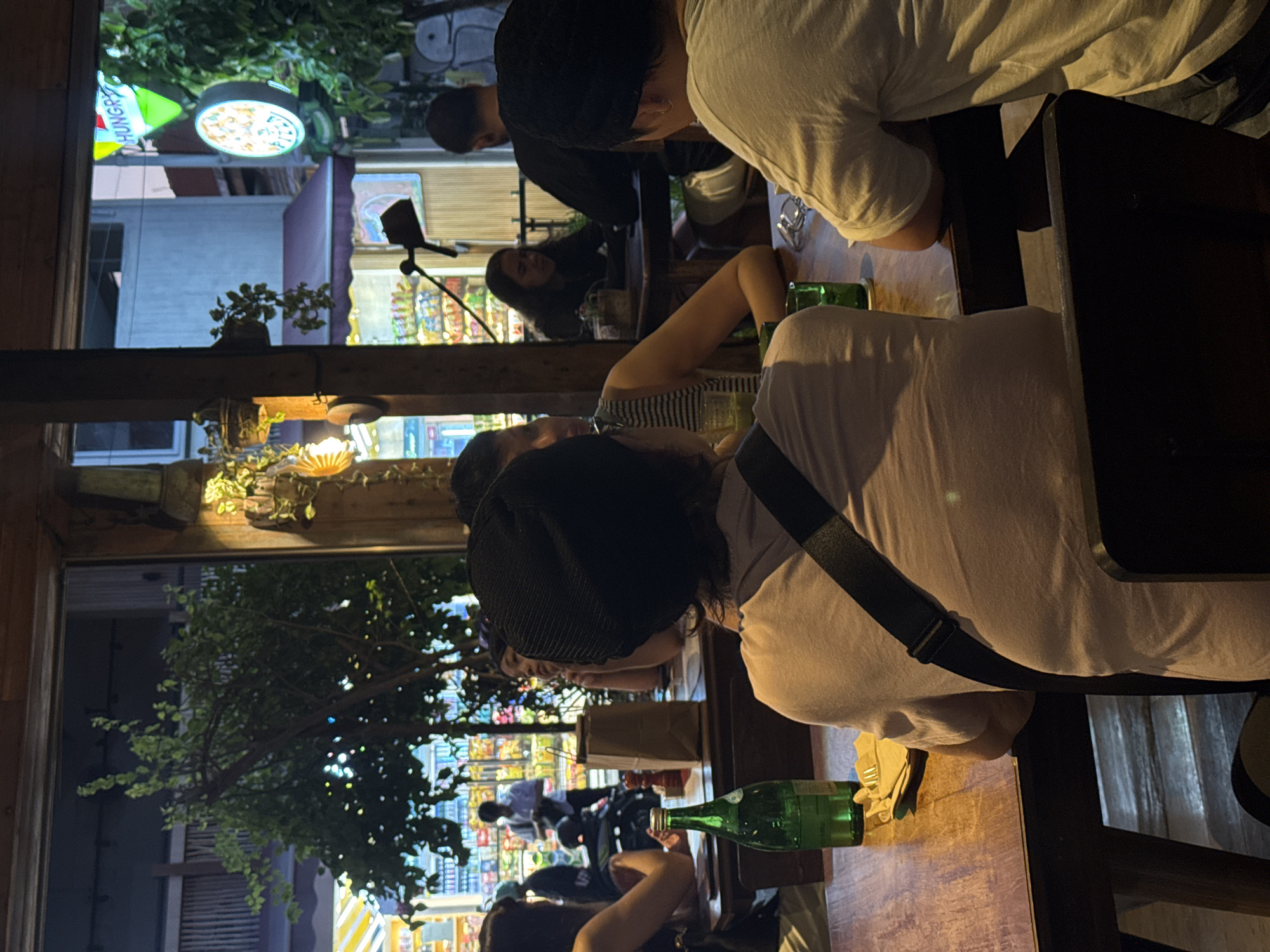 The image shows a group of people sitting at tables in an outdoor dining area of a restaurant in North Kuta, Indonesia. The scene is illuminated by artificial lights, creating a warm and inviting ambiance. In the foreground, a person wearing a black beanie and a light-colored t-shirt is seated at a wooden table. In front of them is a green glass bottle and a plate with utensils. Other diners are visible in the background, some engaged in conversation. Beyond the dining area, a street scene is visible with shops and neon signs. One sign reads "HUNGRY," and another displays a circular image of a pizza, suggesting the establishment may serve pizza or other Italian-inspired cuisine. The overall impression is a lively and social gathering at a restaurant.